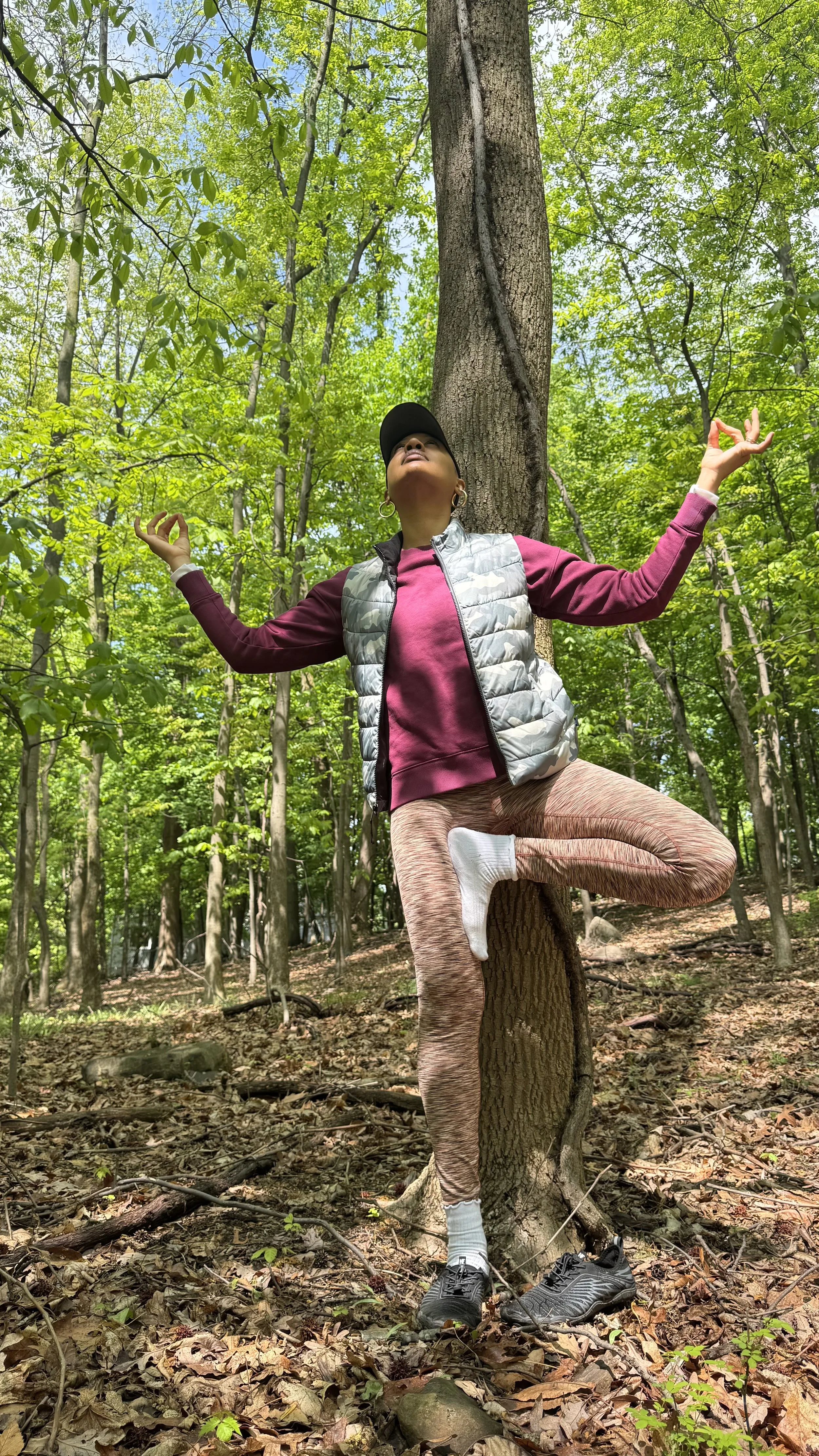 Tiffany in a forest in tree pose with arms in a meditative gesture, leaning against a tree trunk, wearing athletic clothing and sneakers.