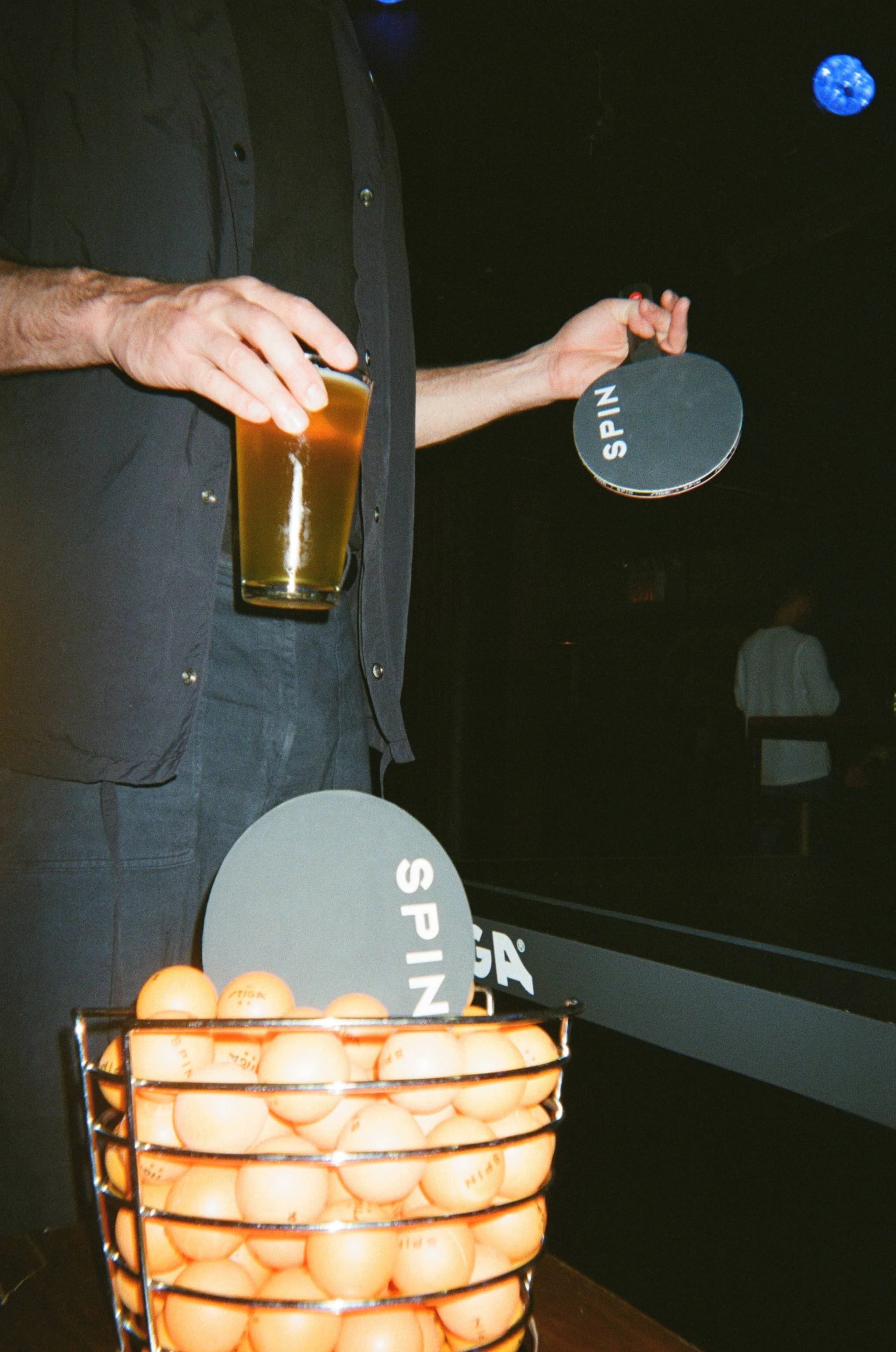 Hand holding a Spin ping pong paddle and beer next to a basket of orange ping pong balls at a Toronto social club