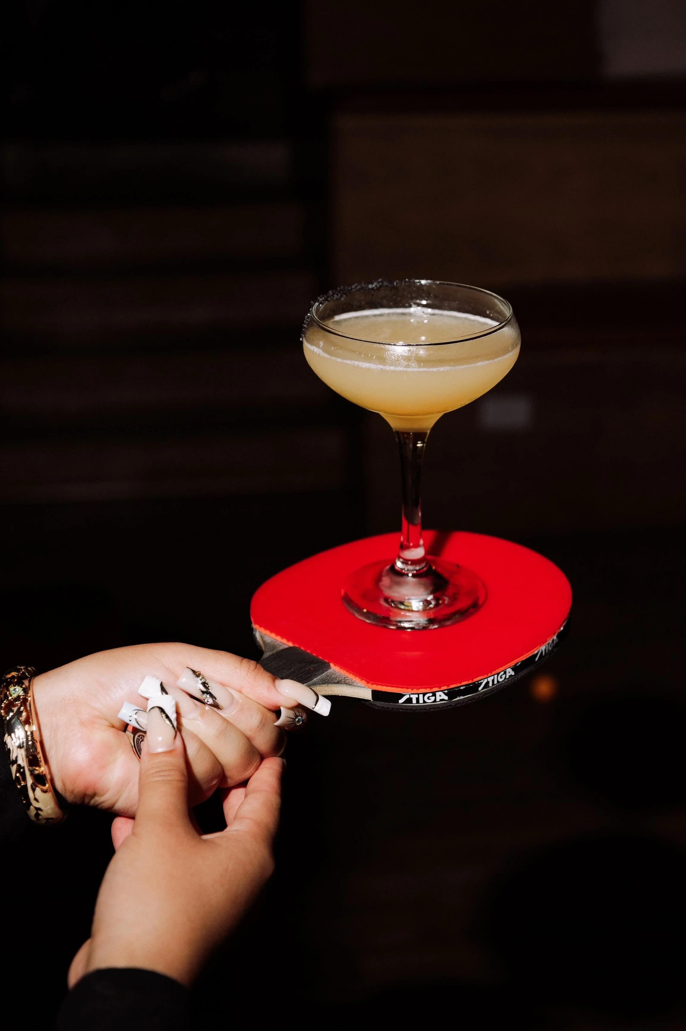 Cocktail in a coupe glass balanced on a red Stiga ping pong paddle held by a hand with manicured nails, nightlife content for a Toronto social club