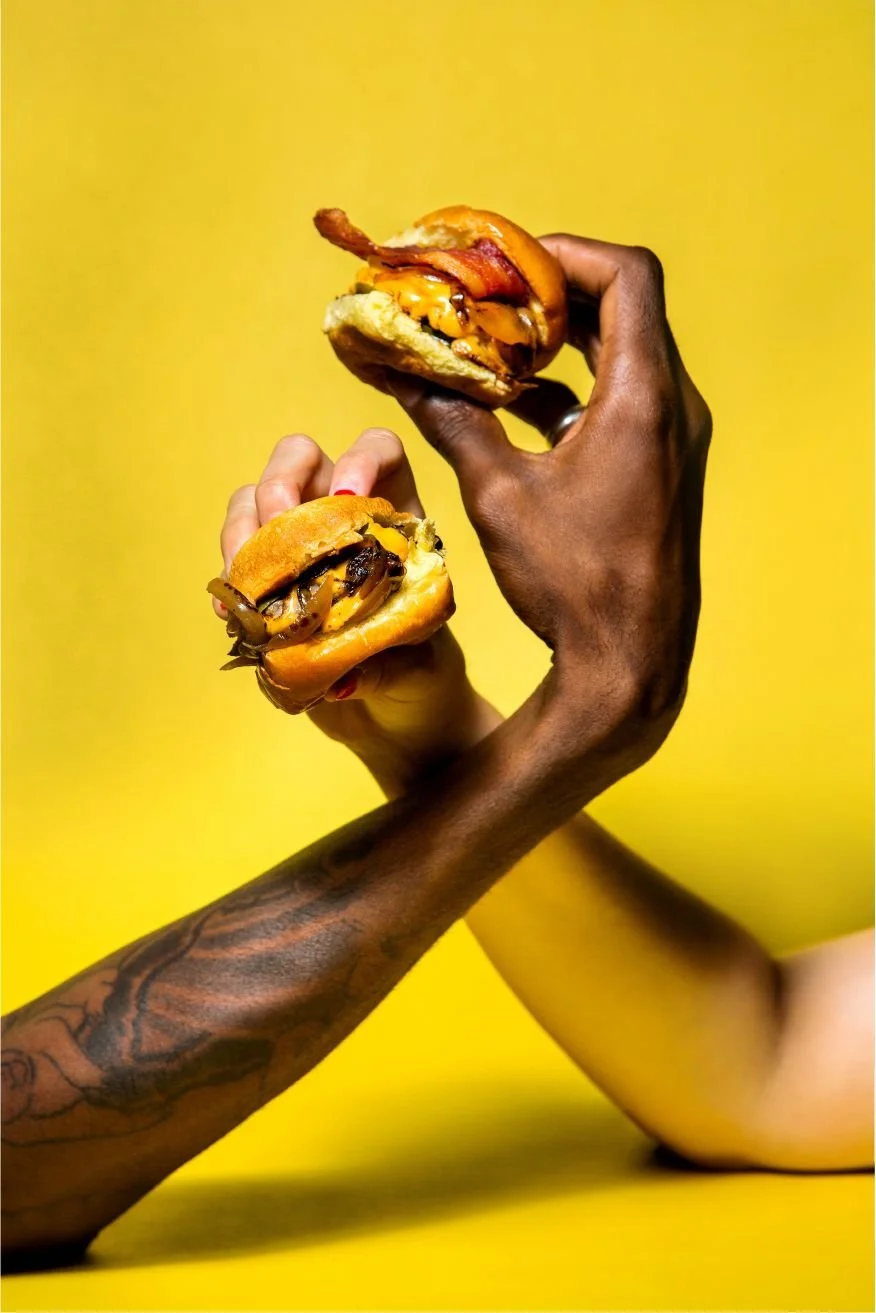 Two hands holding halves of a bitten smash burger slider against a bright yellow background