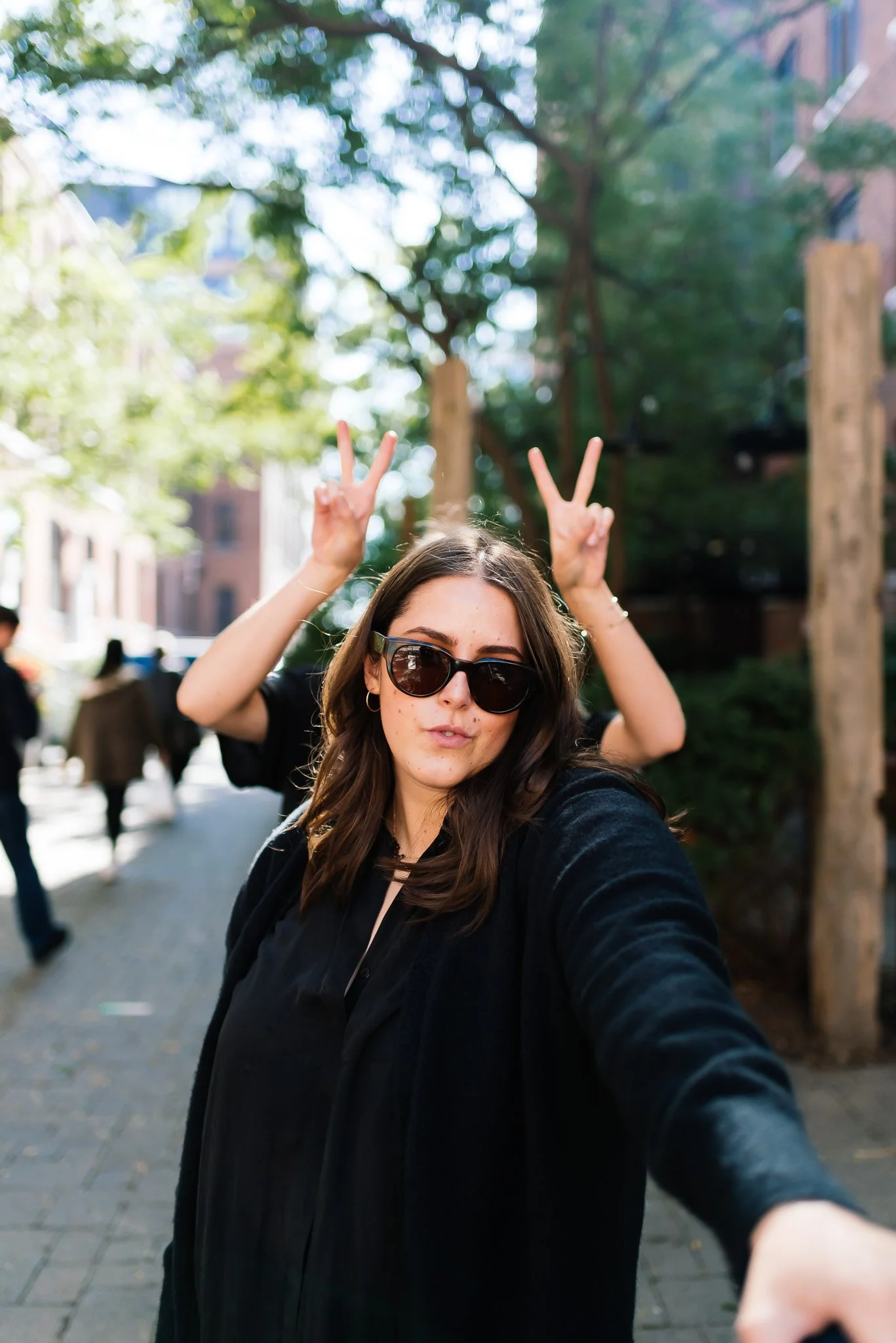 Chloe Castonguay founder of Banquette in sunglasses giving a peace sign with both hands on a tree-lined city sidewalk