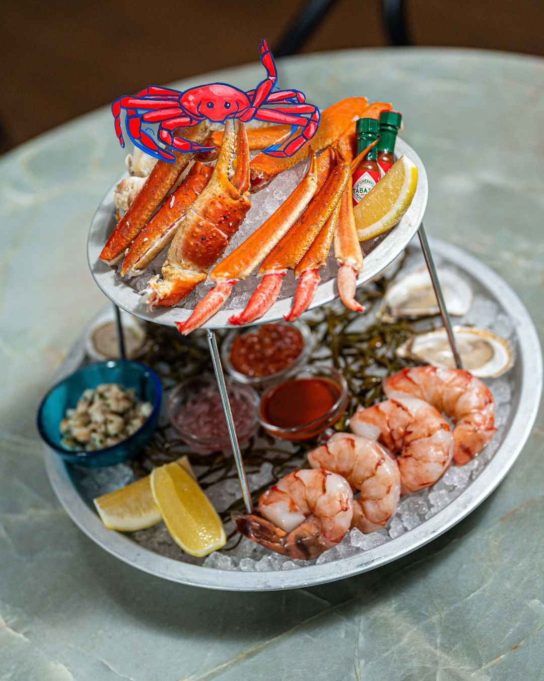 Tiered seafood tower with crab legs, shrimp and oysters on ice with hot sauce and lemon, plus an illustrated red crab graphic overlay, hospitality content for a Toronto restaurant