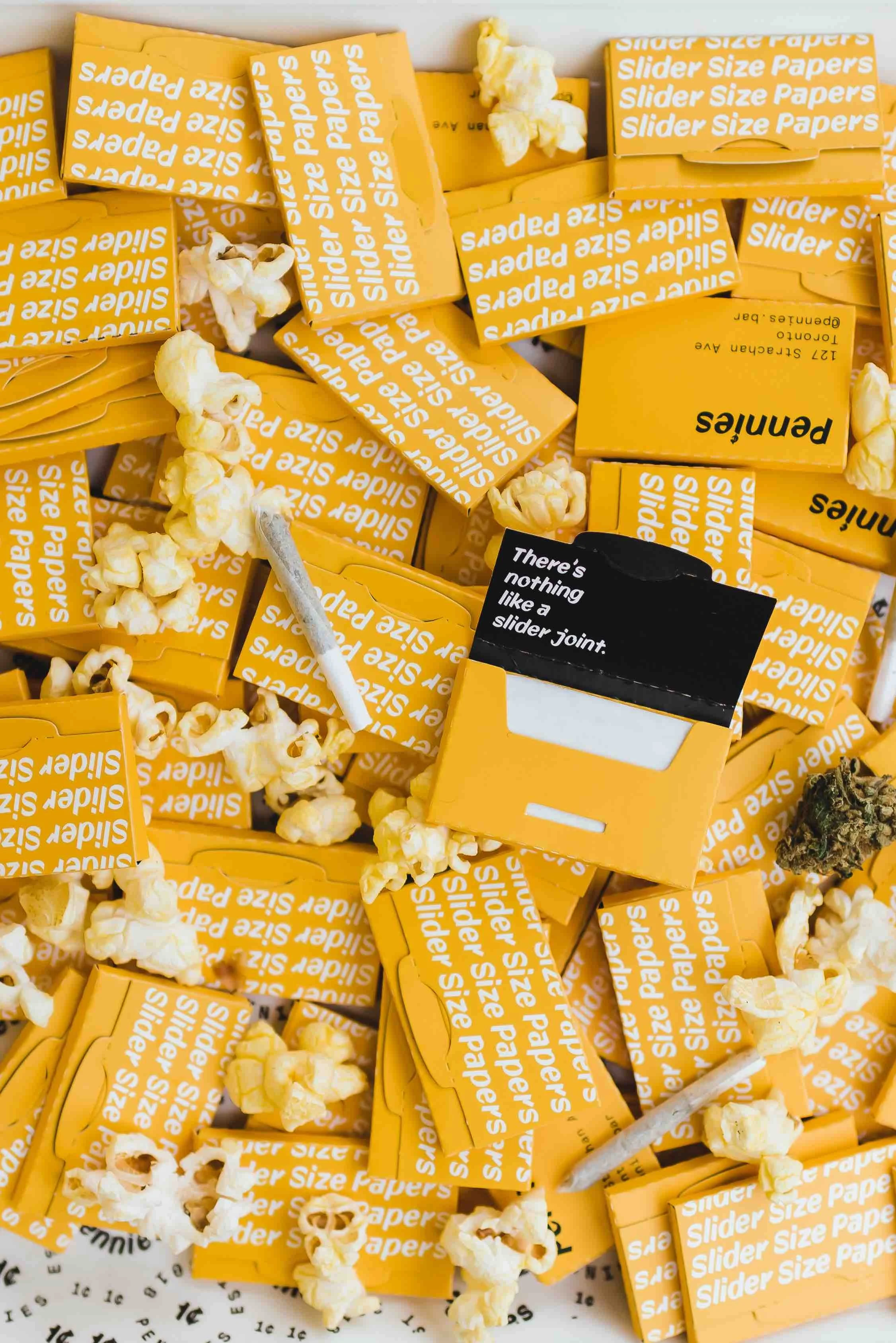 Pile of yellow Pennies branded slider size rolling papers scattered with popcorn and a rolled joint
