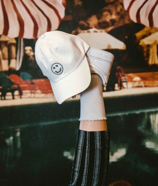 White baseball cap with Pennies logo and white sneaker against a poolside mural backdrop, branded merch content for a Toronto hospitality business