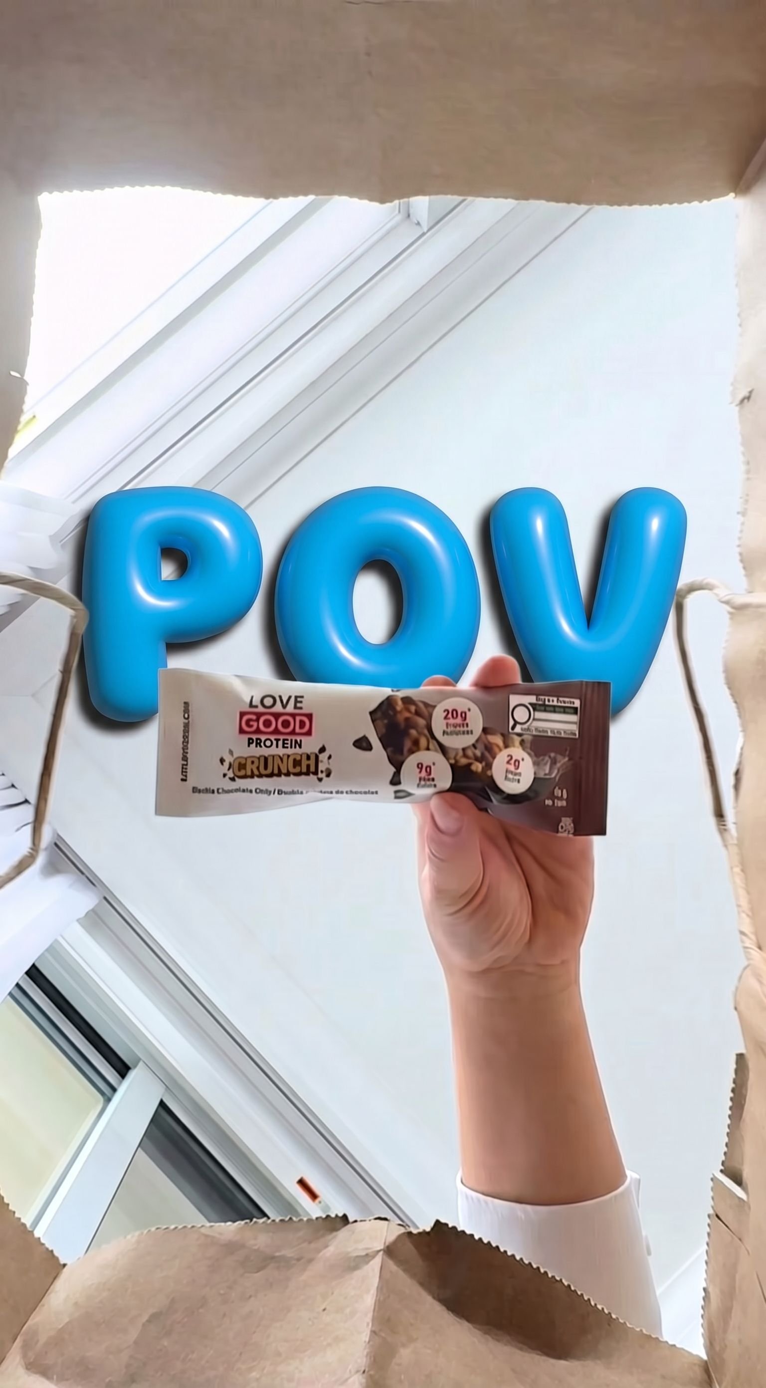 Hand holding a Love Good Protein Crunch chocolate bar in front of a doorway with inflatable blue POV letters