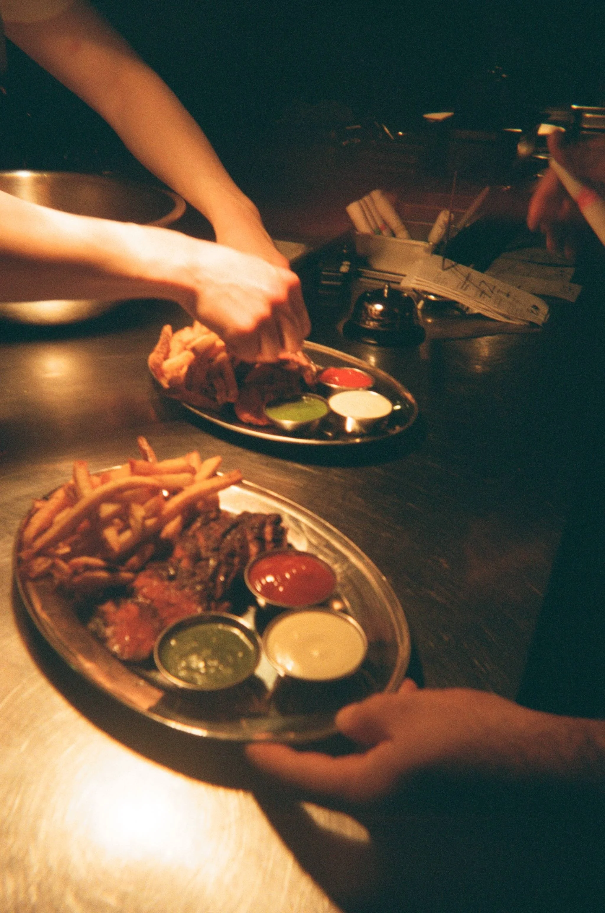 Overhead shot of two hands passing metal trays of fries, wings and dipping sauces in a warmly lit bar, communications content for a Toronto hospitality business