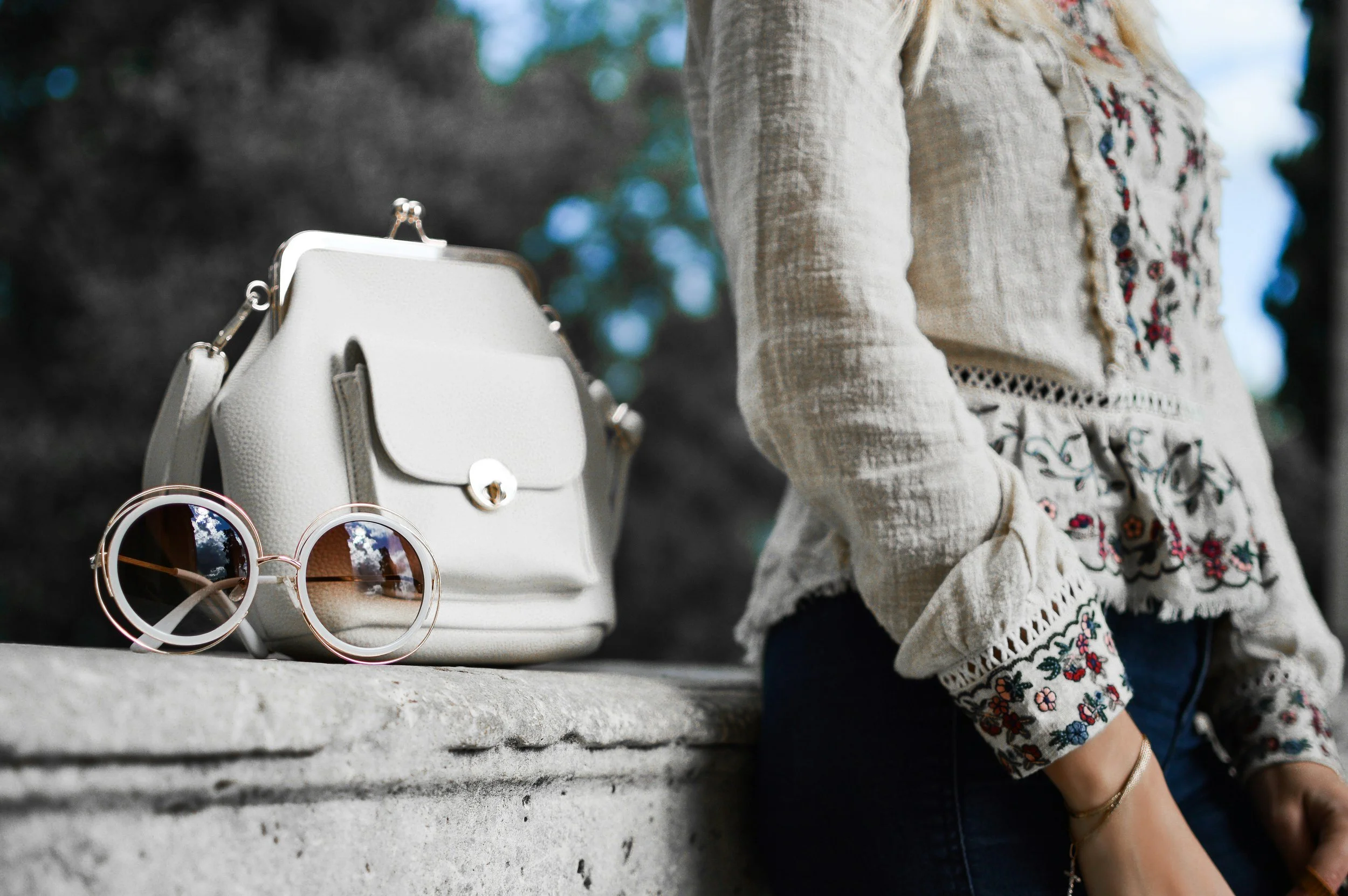 A person wearing a light-colored embroidered blouse with floral patterns, standing outdoors near a stone ledge. On the ledge, there is a white handbag with a gold clasp and two pairs of round sunglasses with reflective lenses.
