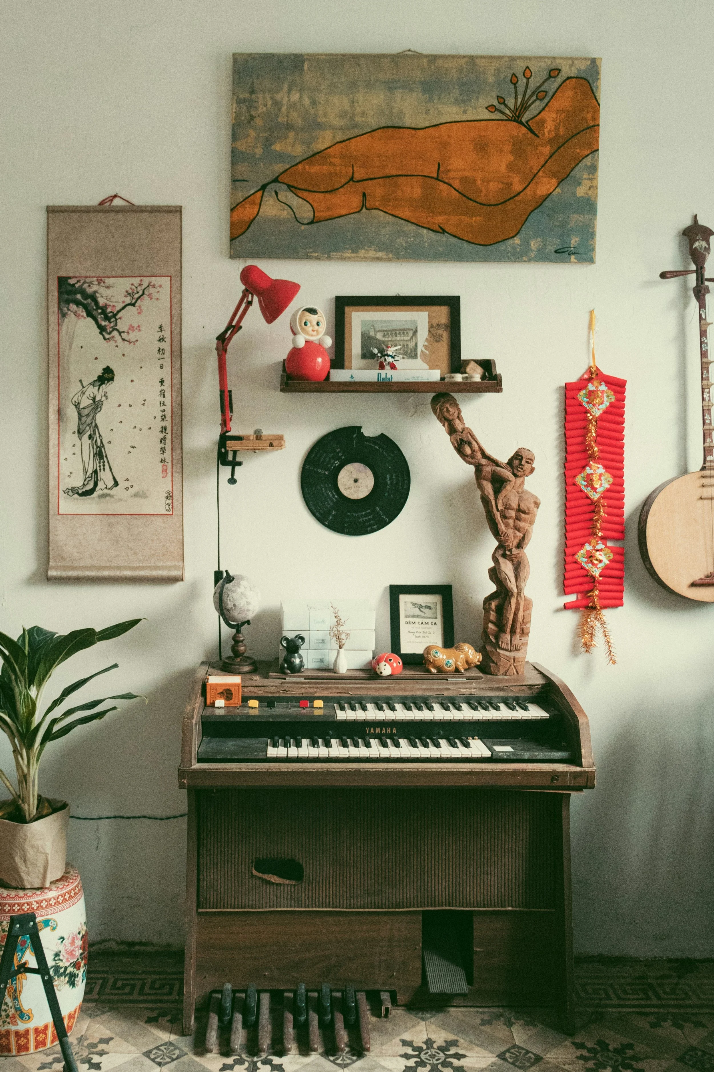 A living room corner with a Yamaha keyboard on the floor, a potted plant on the left, wall decorations including a large painting of a hand holding a flower, a smaller framed picture, a hanging scroll with a woman and cherry blossoms, a red lantern decoration, and a carved wooden sculpture of a human figure.