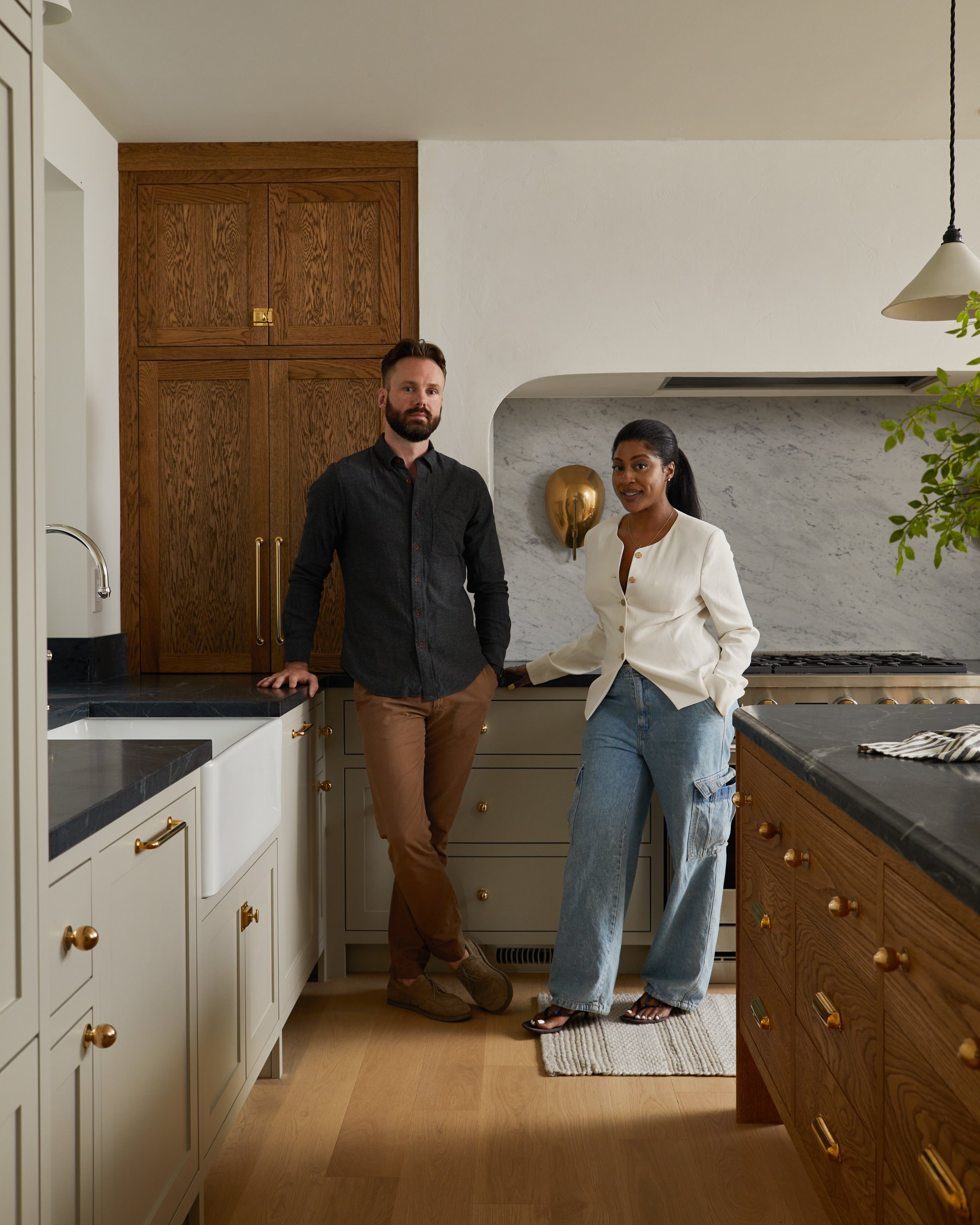 A man and a woman standing in a kitchen, looking at the camera. The man has a beard, wears a black shirt and brown pants, and leans against a white cabinet. The woman has dark hair pulled back, wears a cream blazer and blue jeans, and is smiling. The kitchen has wooden cabinets, a marble backsplash, and a black countertop.