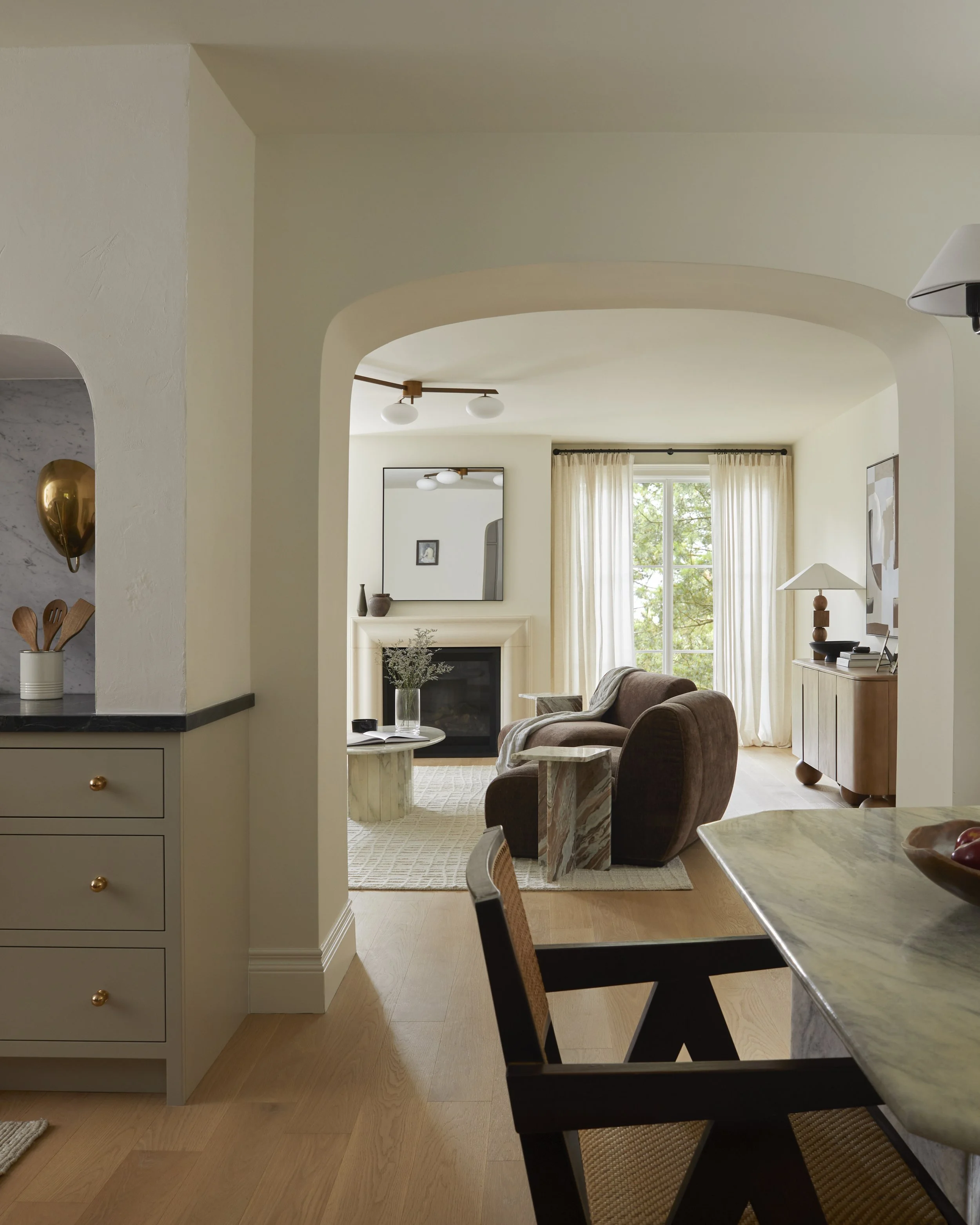 The image shows a view from a dining area into a living room. The dining table has a marble top with a black chair in the foreground. In the living room, there is a sofa with a blanket, a round stone-like coffee table, a fireplace with decorative vases above it, and large windows with light curtains revealing trees outside. The ceiling has two modern globe lights, and part of a kitchen island with a ceramic utensil holder is visible on the left.