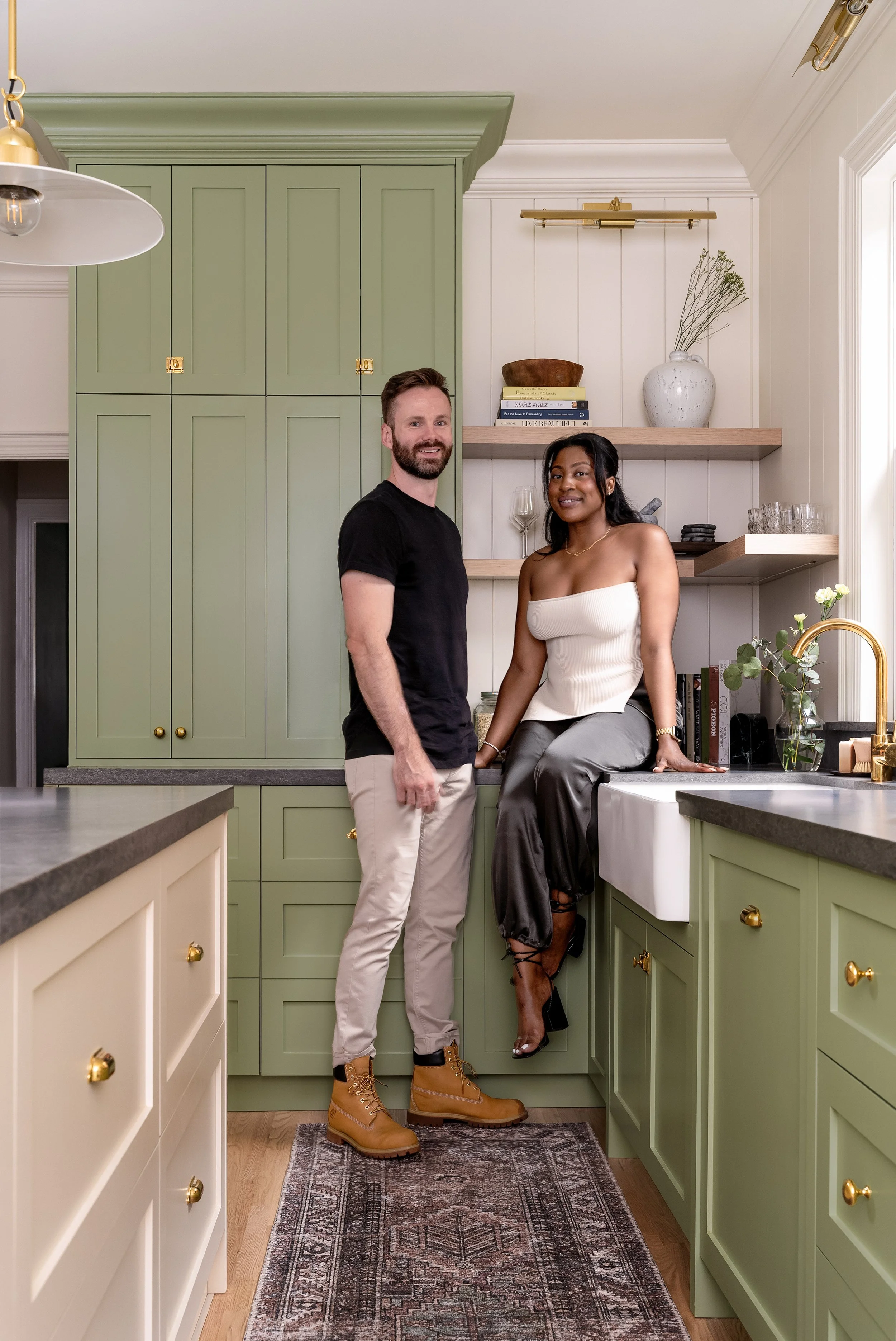 A man and woman in a kitchen with green and cream cabinets, a wooden shelf with books and decor, a vase with greenery, and a patterned rug. The woman is sitting on the counter, and the man is standing beside her.