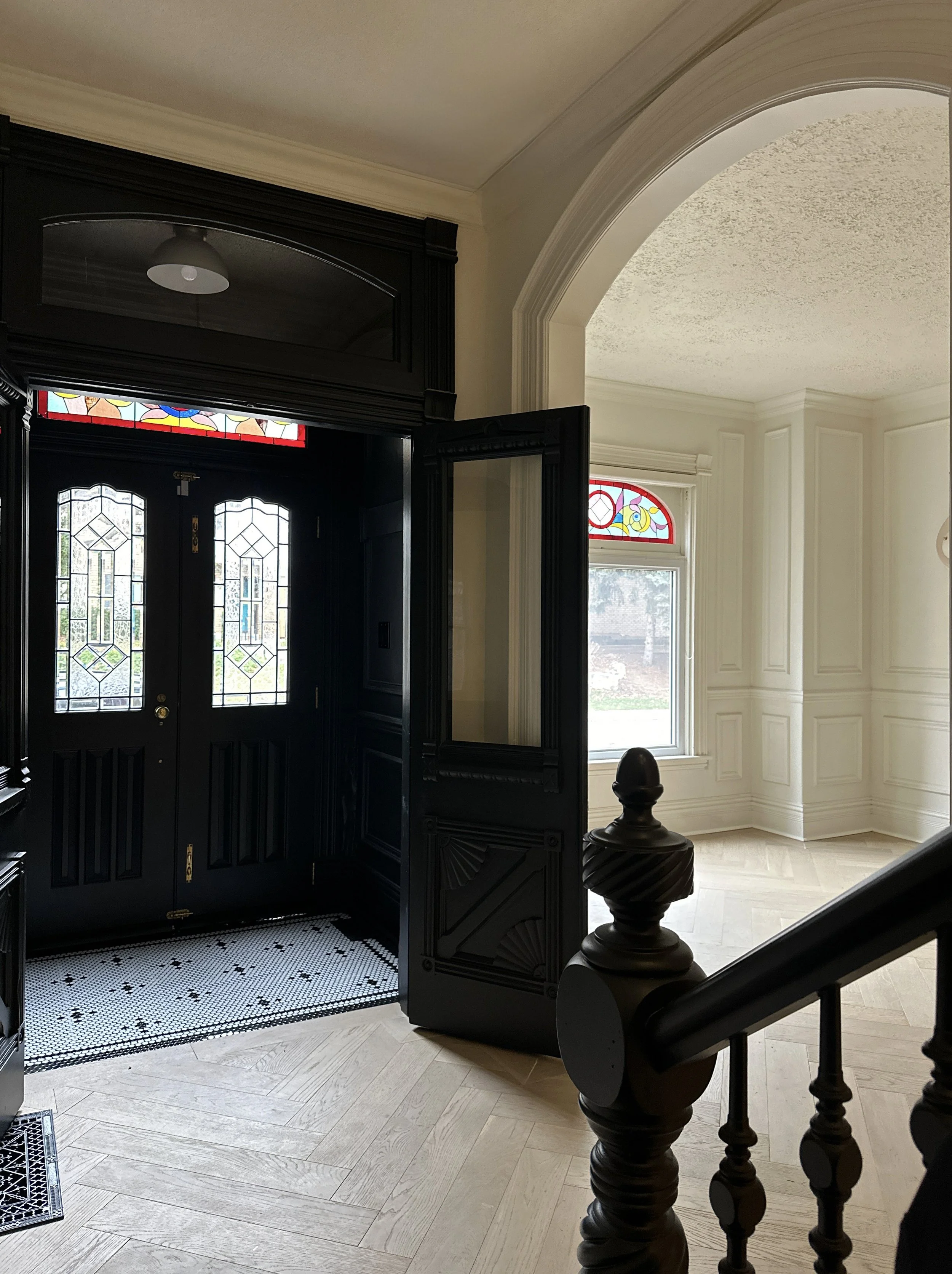 View of an elegant house entrance with black double doors with stained glass windows, an intricate black door frame, a black wall mirror, and a staircase with wooden handrails. Bright natural light fills the space through large windows, and colorful stained glass accents are visible.