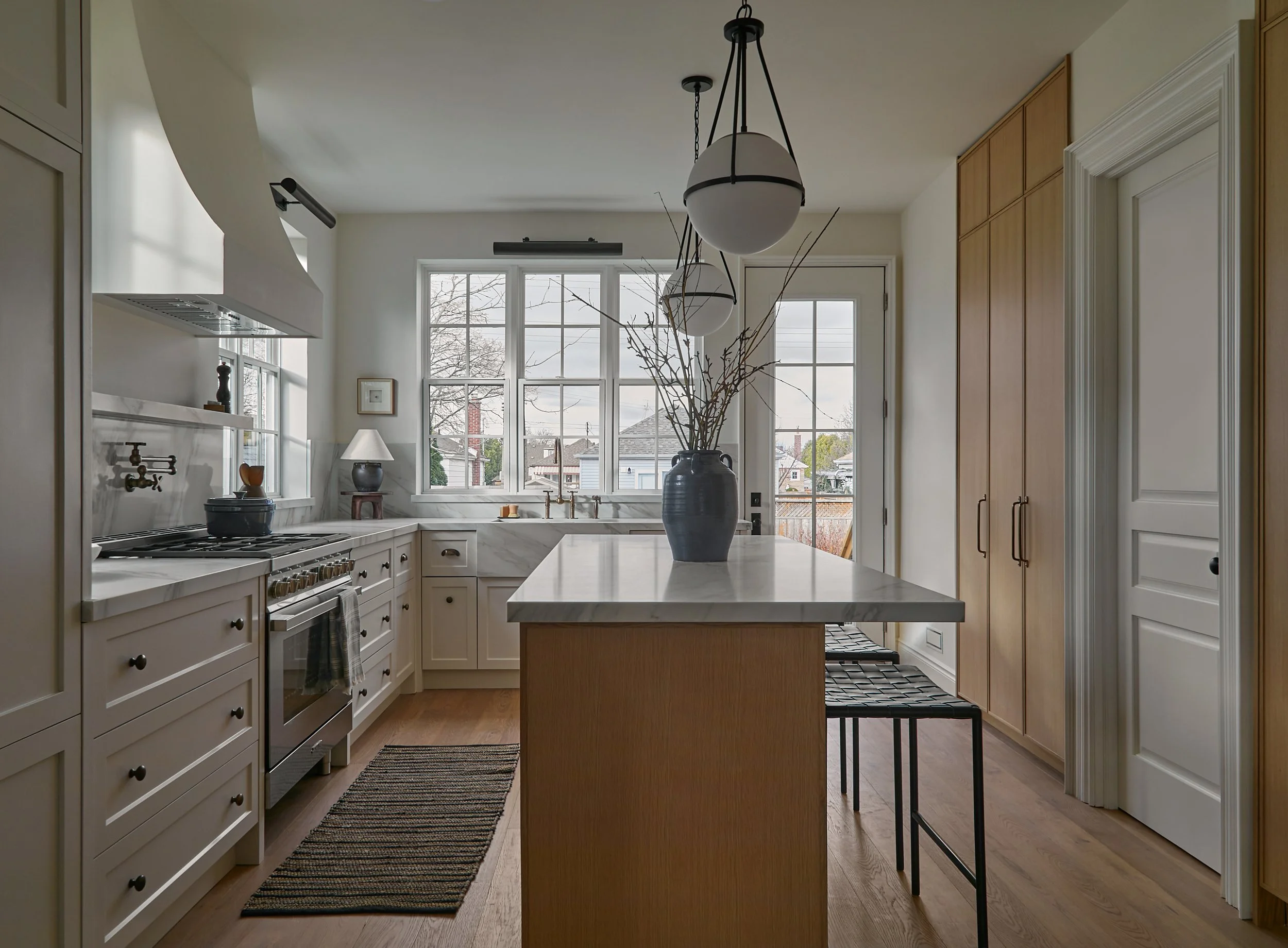 View of a modern kitchen with white cabinetry, marble countertops, a large island, and wooden cabinets. Two large pendant lights hang above the island, and a large window overlooks a neighborhood with trees and houses. Decor includes a big vase with branches and small framed pictures.