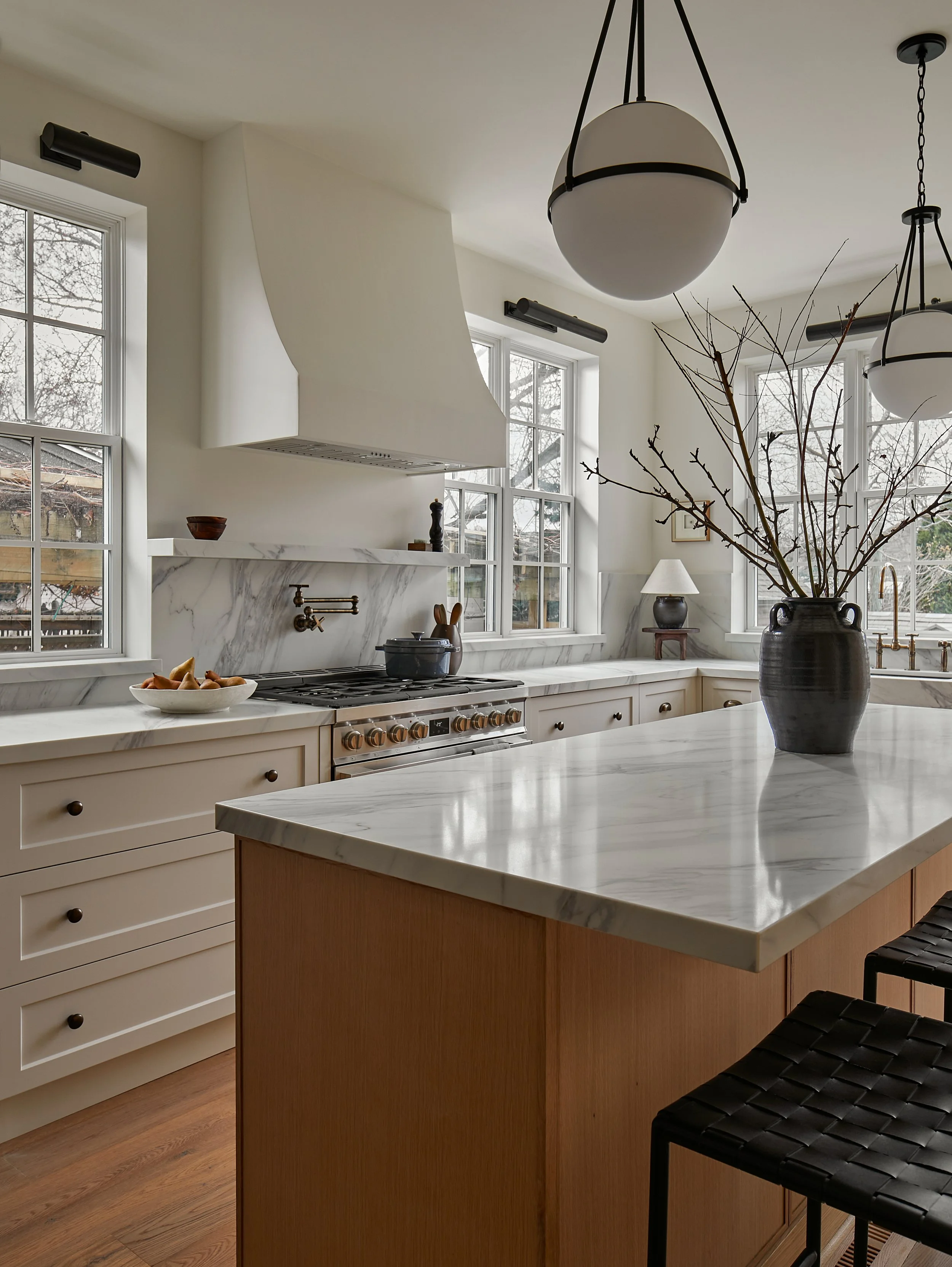 A modern kitchen with white and wood cabinets, marble countertops, and a large island. There are three large white pendant lights hanging from the ceiling, and a black wicker chair at the island. The kitchen has multiple windows allowing natural light to illuminate the space.