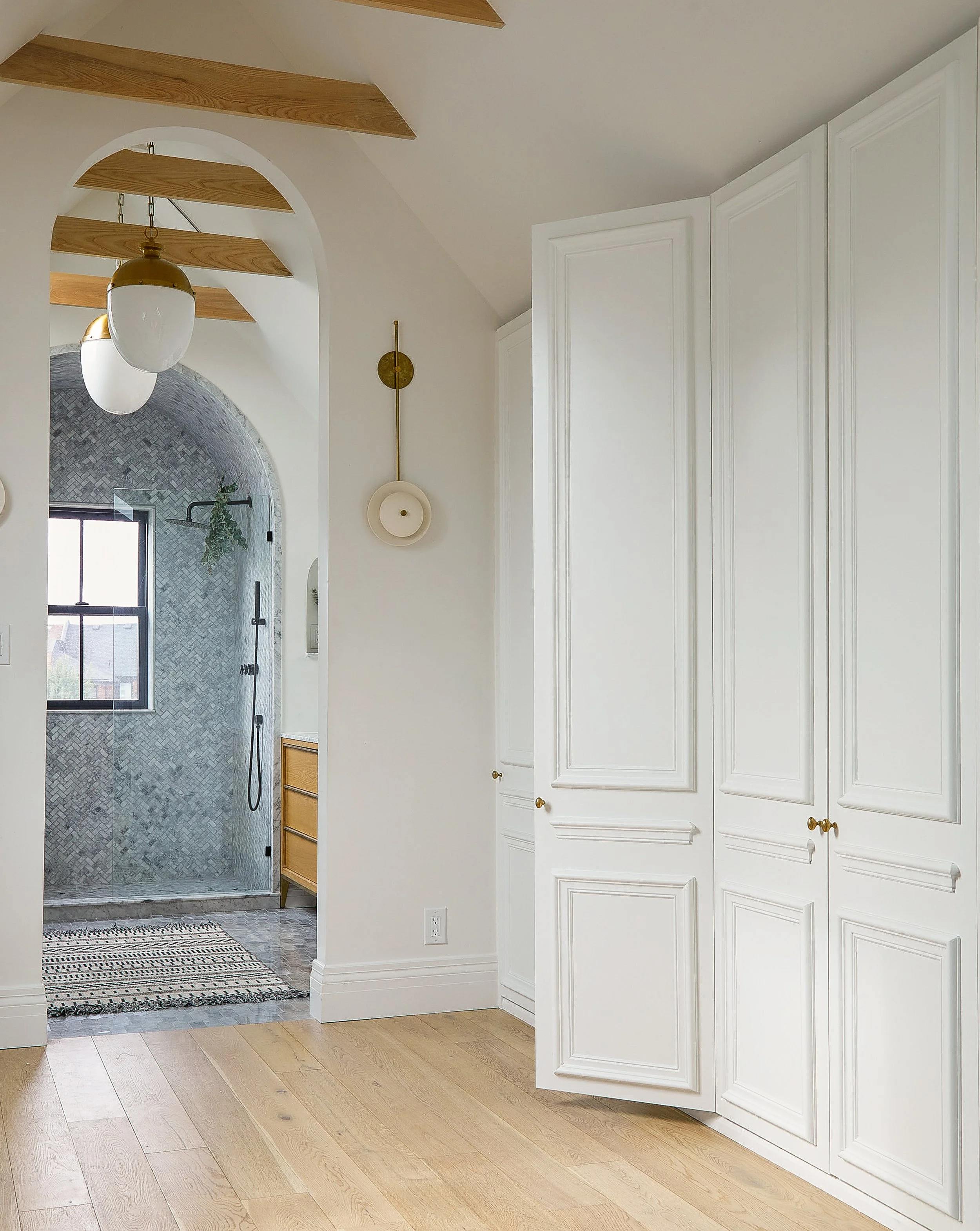 Interior view of a modern kitchen with white cabinets, hardwood floors, and a doorway leading to a shower area with gray mosaic tile walls and a window. A decorative wall light and a black and white rug are visible.