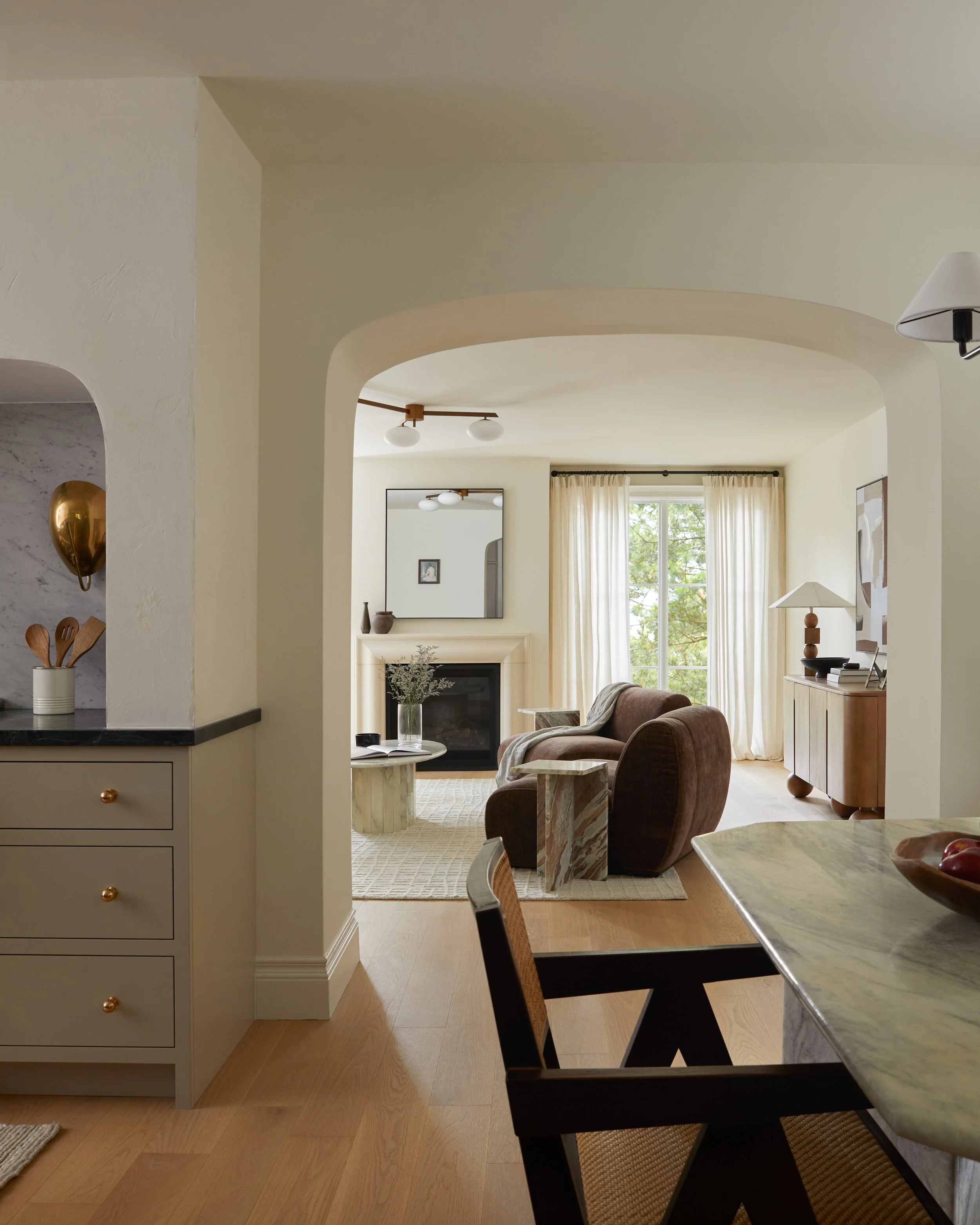 A view from inside a house showing the living and dining areas. The foreground has a part of a kitchen counter with white drawers and gold knobs, a container with wooden utensils, and a part of a chair. Beyond the archway, there is a cozy living room with a fireplace, a mirror above it, a small table, and a cozy armchair. Large windows with curtains let in natural light, and a chandelier with round white globes hangs from the ceiling.