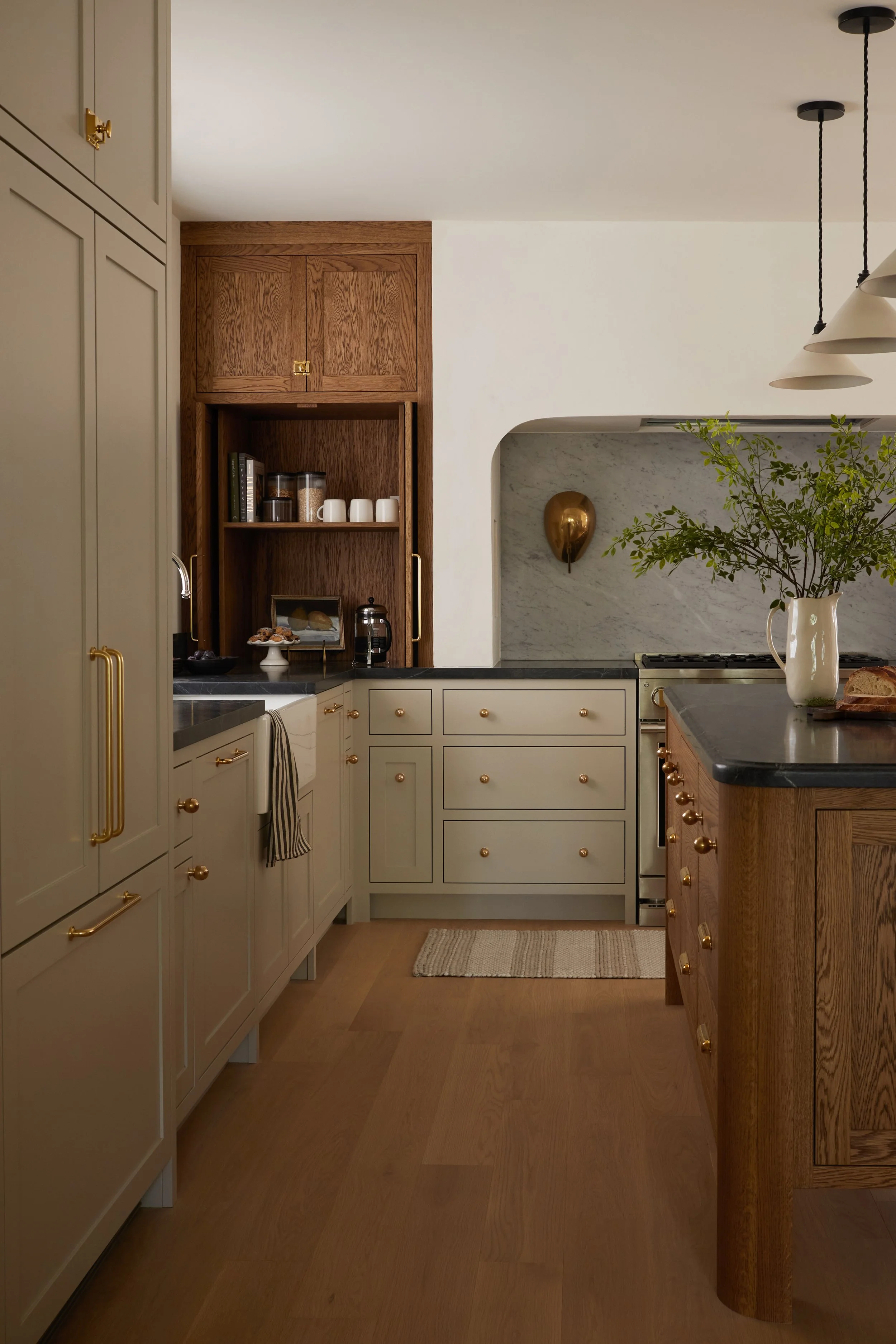 A modern kitchen with beige cabinets, gold handles, and a dark countertop. There is a wooden cabinet section with open shelves holding kitchen jars, mugs, a dessert stand, and a framed photo. A marble backsplash features a gold wall sconce, and a vas