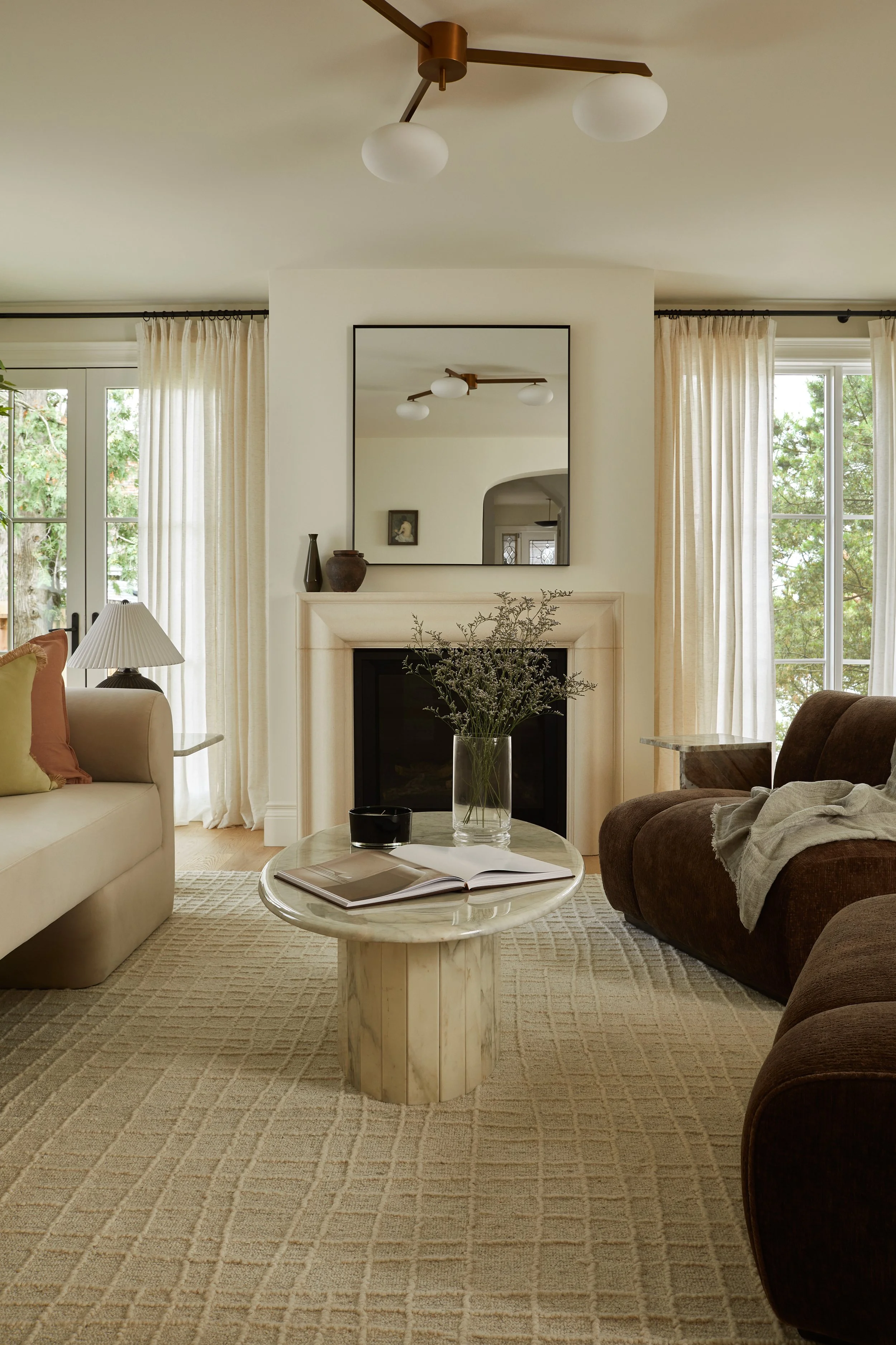 Living room with a cream-colored sofa, a brown armchair, and a marble-top coffee table with a book and a black bowl, positioned on a beige patterned rug. In the background, there is a fireplace with a large rectangular mirror above it, decorative vas