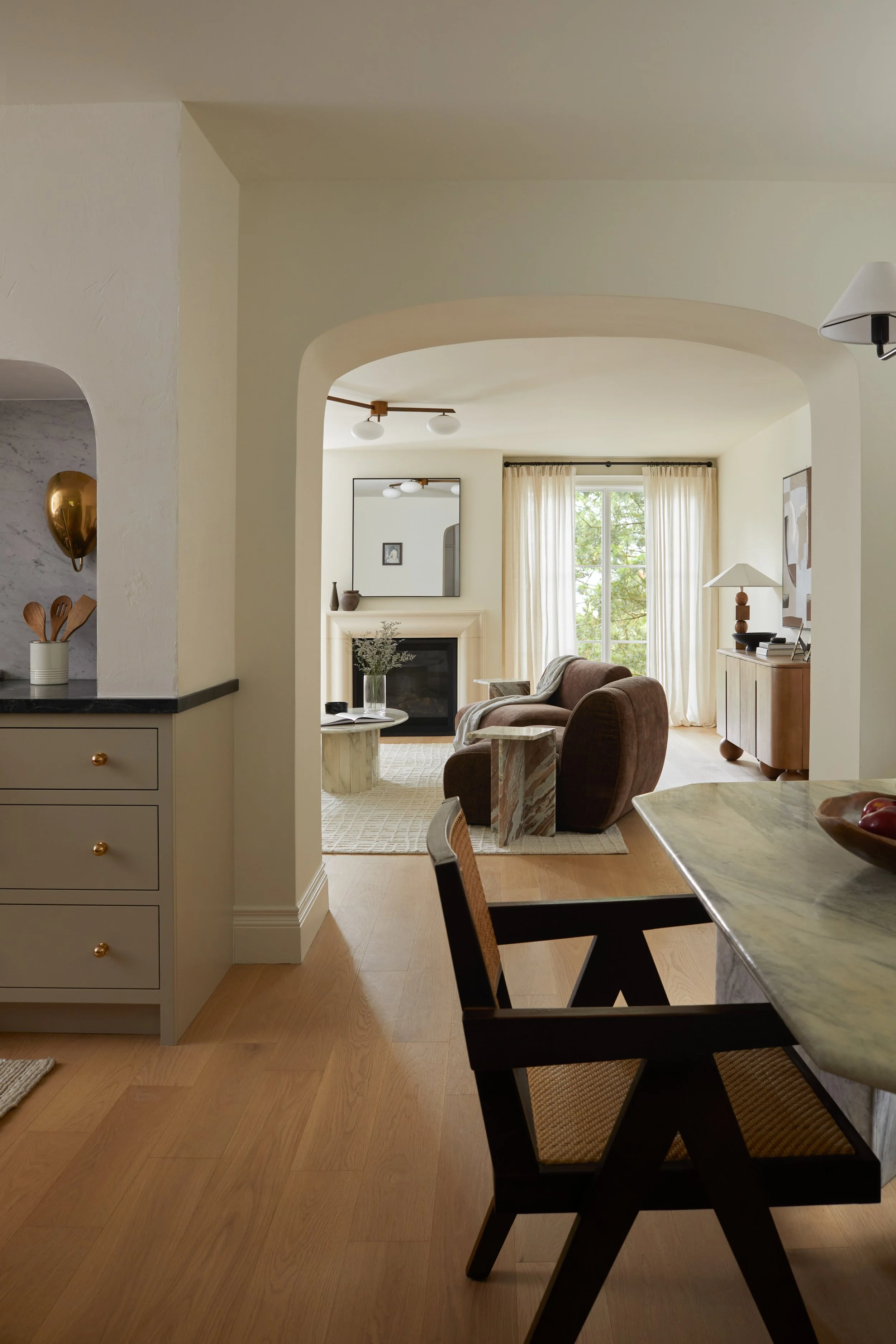 A cozy living room with a large window, beige curtains, a brown armchair, a fireplace with a mirror above, a side table with a lamp, and a dining table with black chairs and a green marble surface.