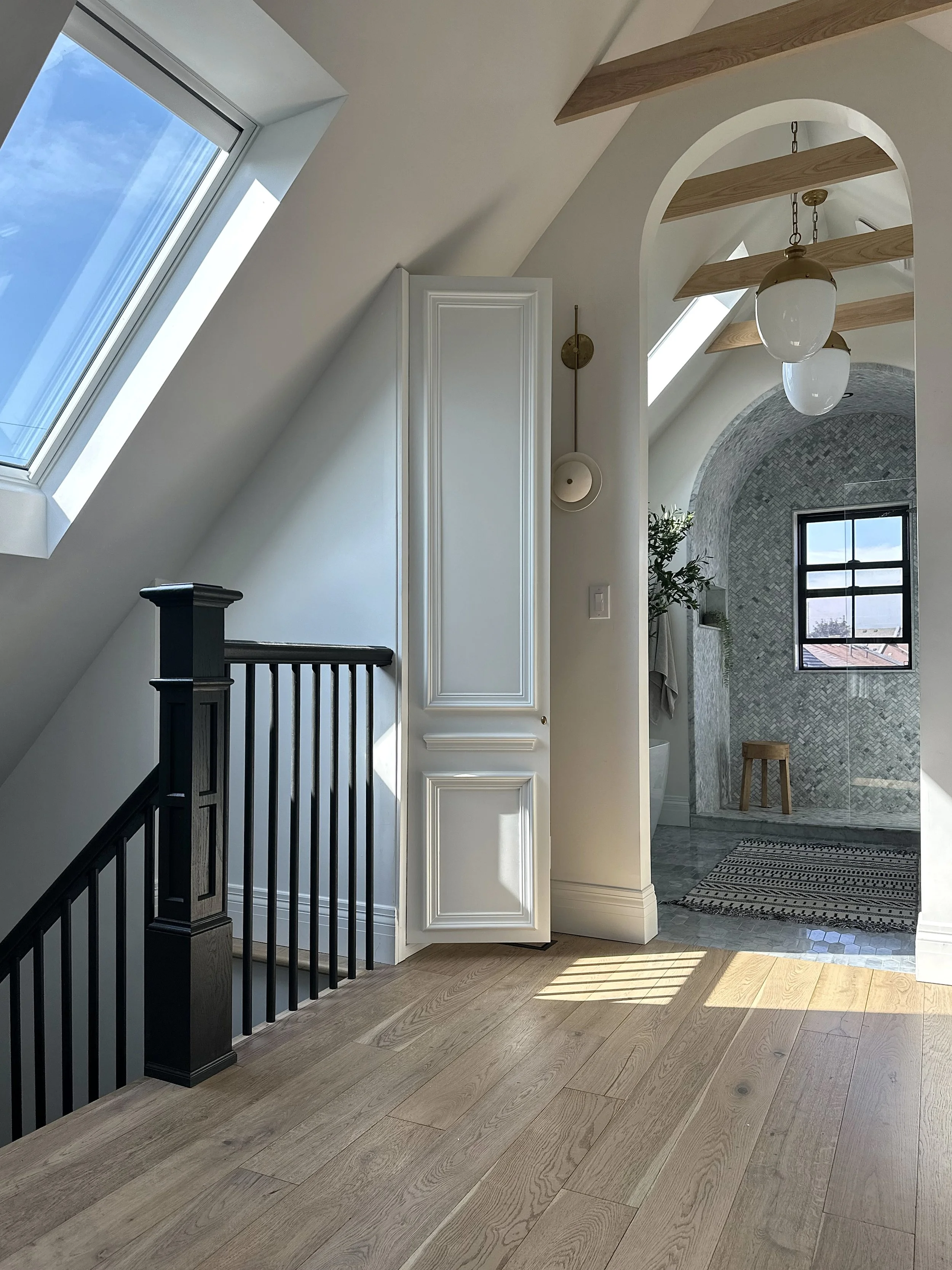 A view of an attic bathroom with a sloped ceiling with skylights, a white cabinet, a wooden staircase railing, a rustic pendant light, a window with a view, and a tiled shower area with a small wooden stool and a patterned rug on the wooden floor.