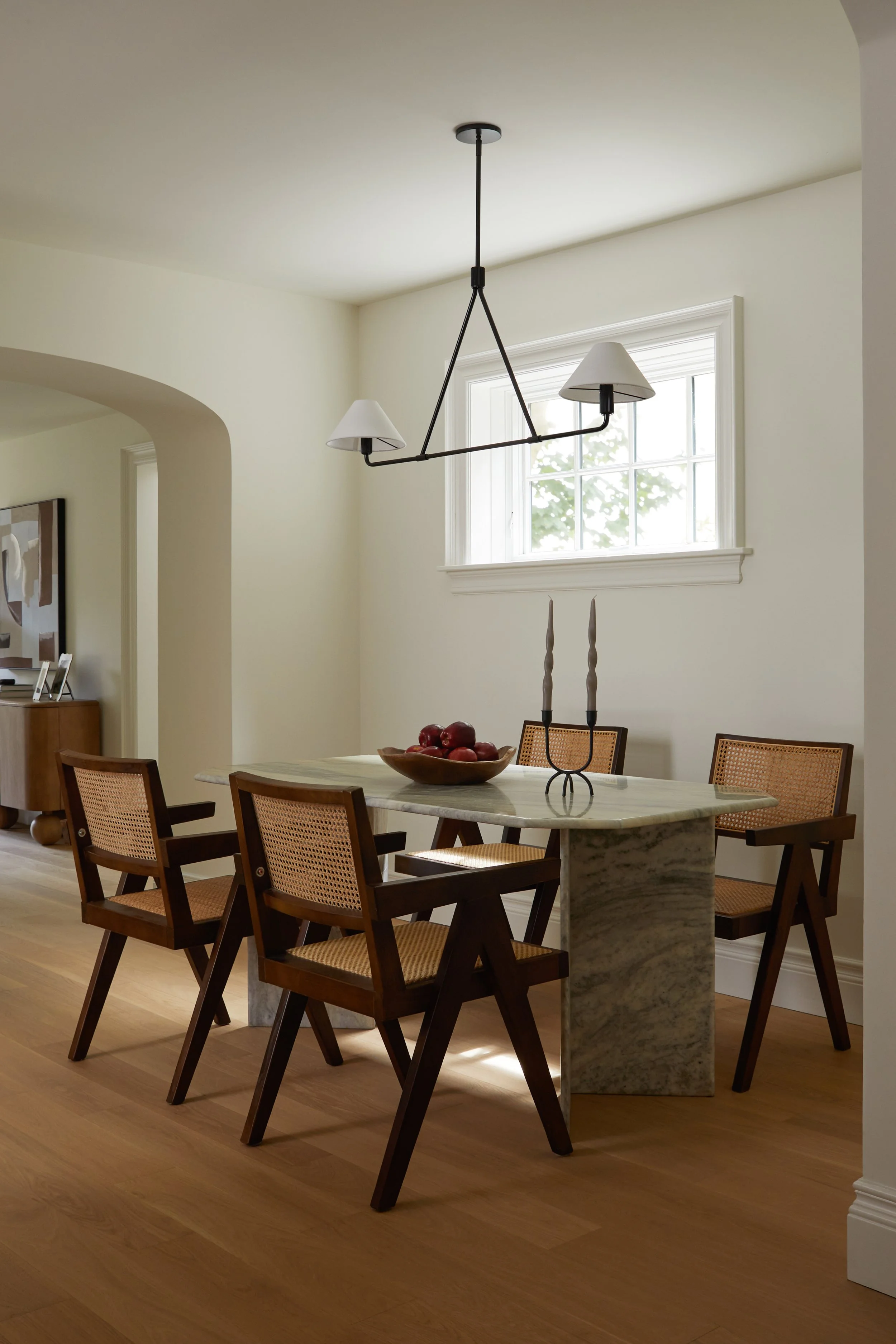 A dining room with a marble table, six wooden chairs with woven cane backs, a bowl of apples on the table, a modern black chandelier with white lampshades, and a window with white trim letting in natural light.