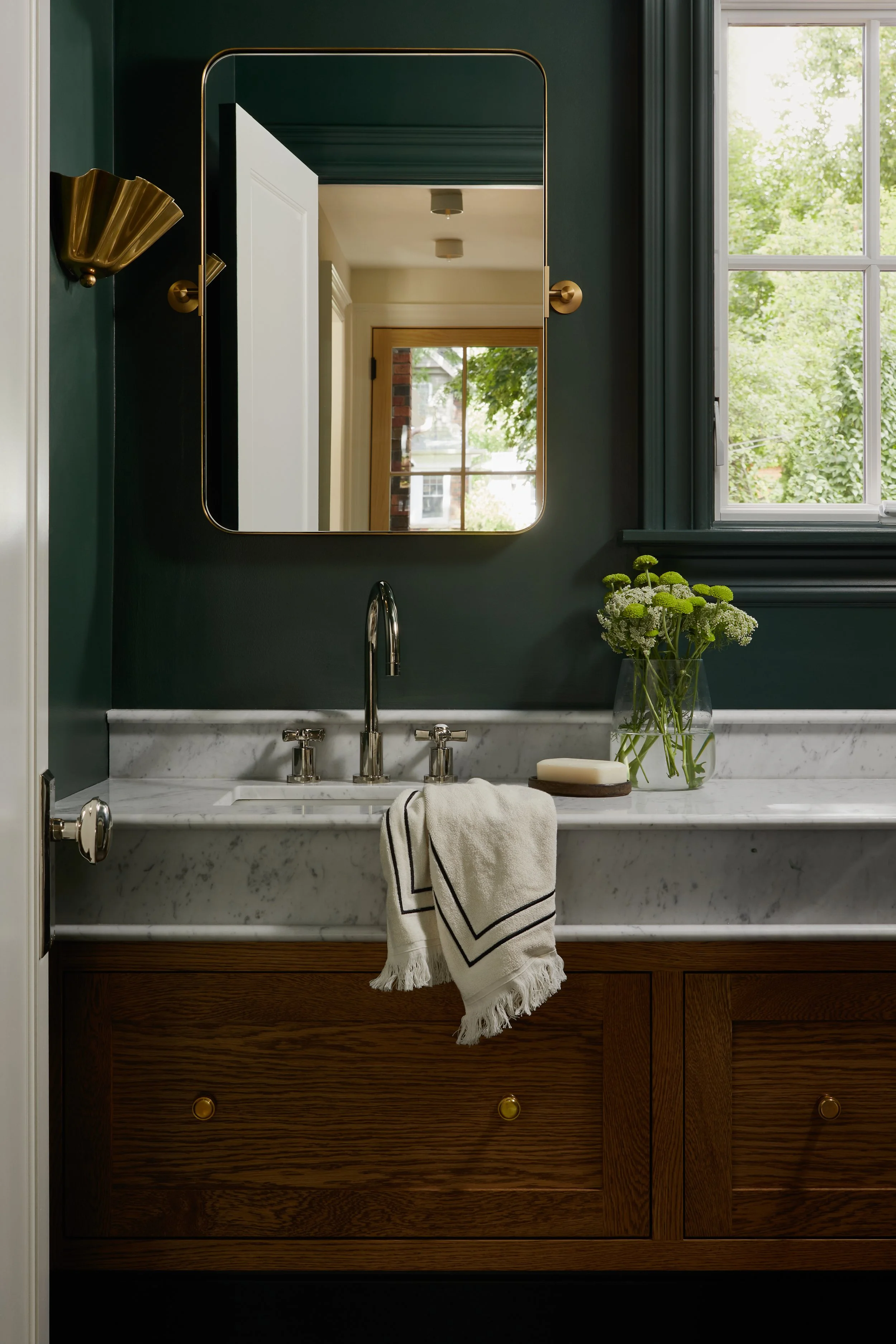 Bathroom vanity with a marble countertop, wooden cabinet, a rectangular mirror, a vase with white and green flowers, and a window with green trees outside.