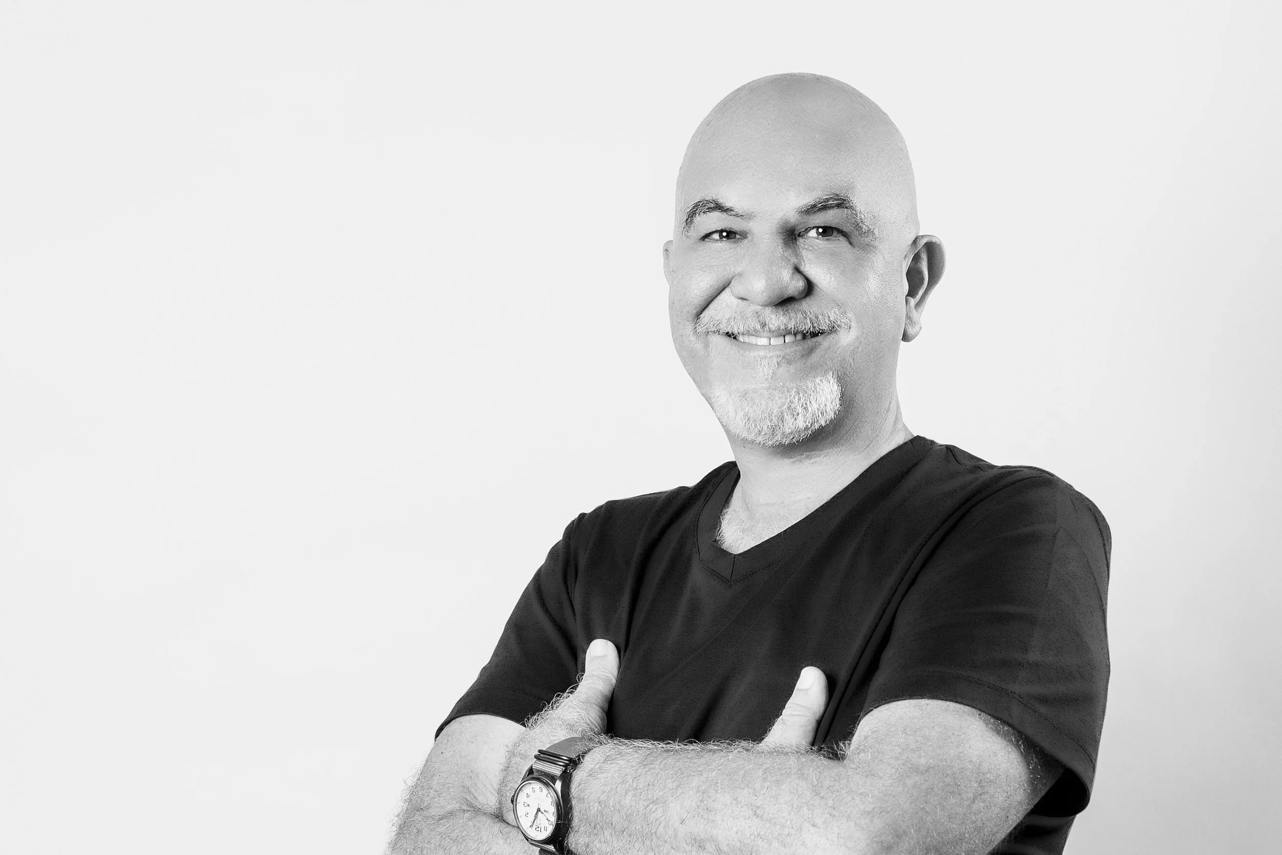 Black and white photo of a middle-aged man smiling, with a bald head, white beard and mustache, wearing a black T-shirt, a watch, and crossing his arms.