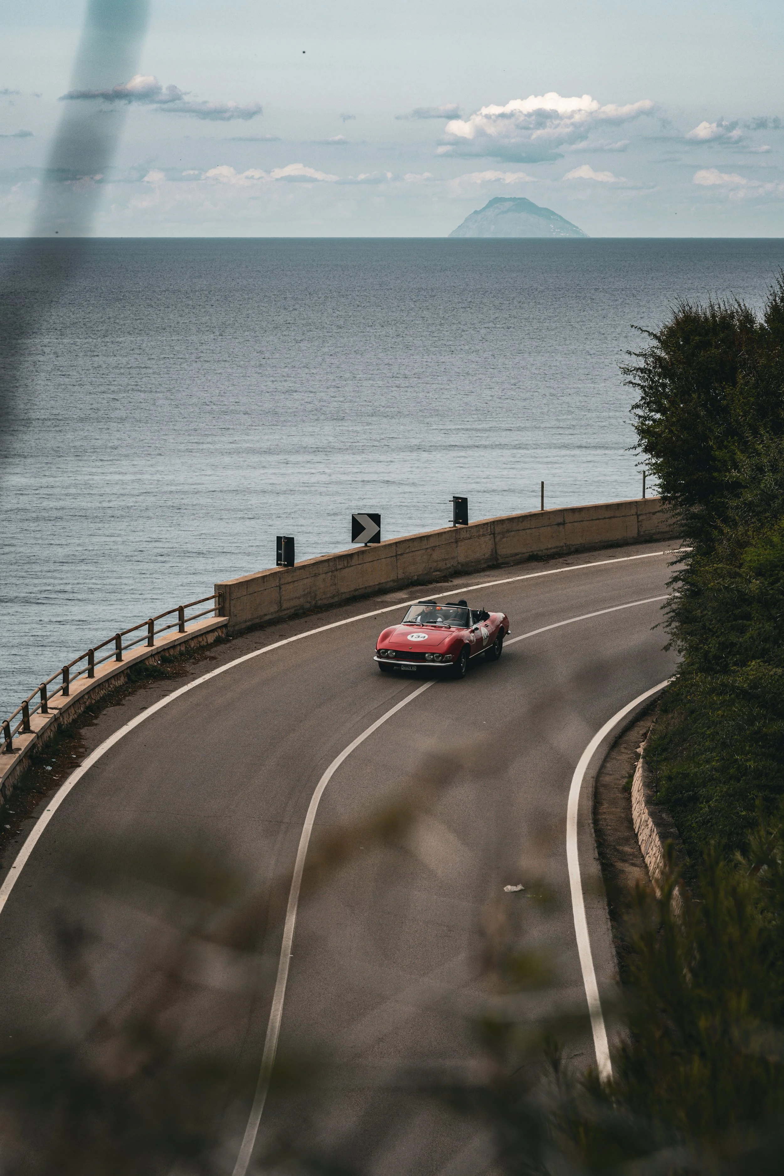 Una strada costiera con un'auto sportiva rossa che percorre una curva, in primo piano ci sono foglie di alberi e una barriera di sicurezza, sullo sfondo un mare con un'isola.