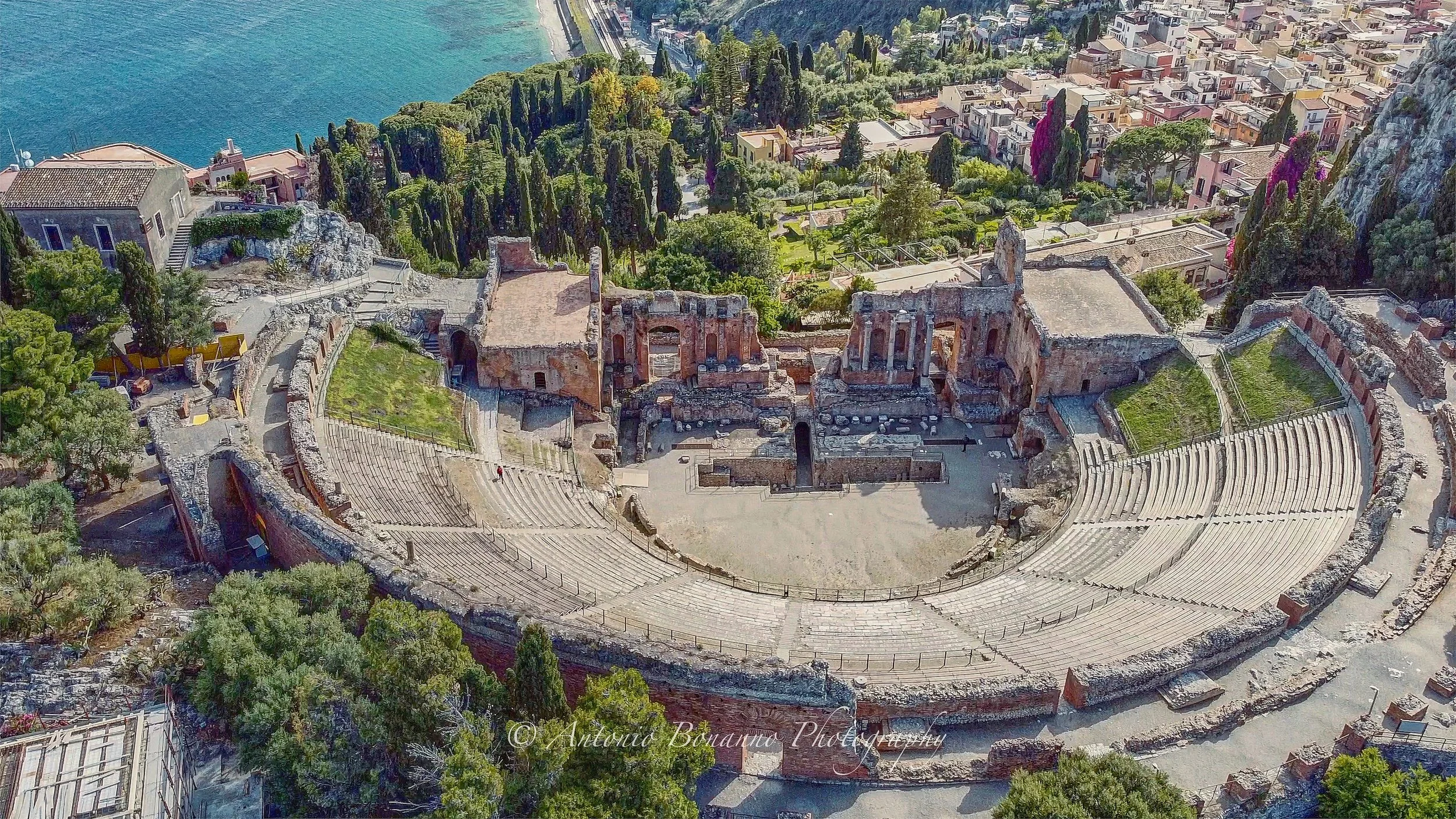 Scorcio aereo di un antico teatro romano in riva al mare con un paesaggio urbano e natura circostante.