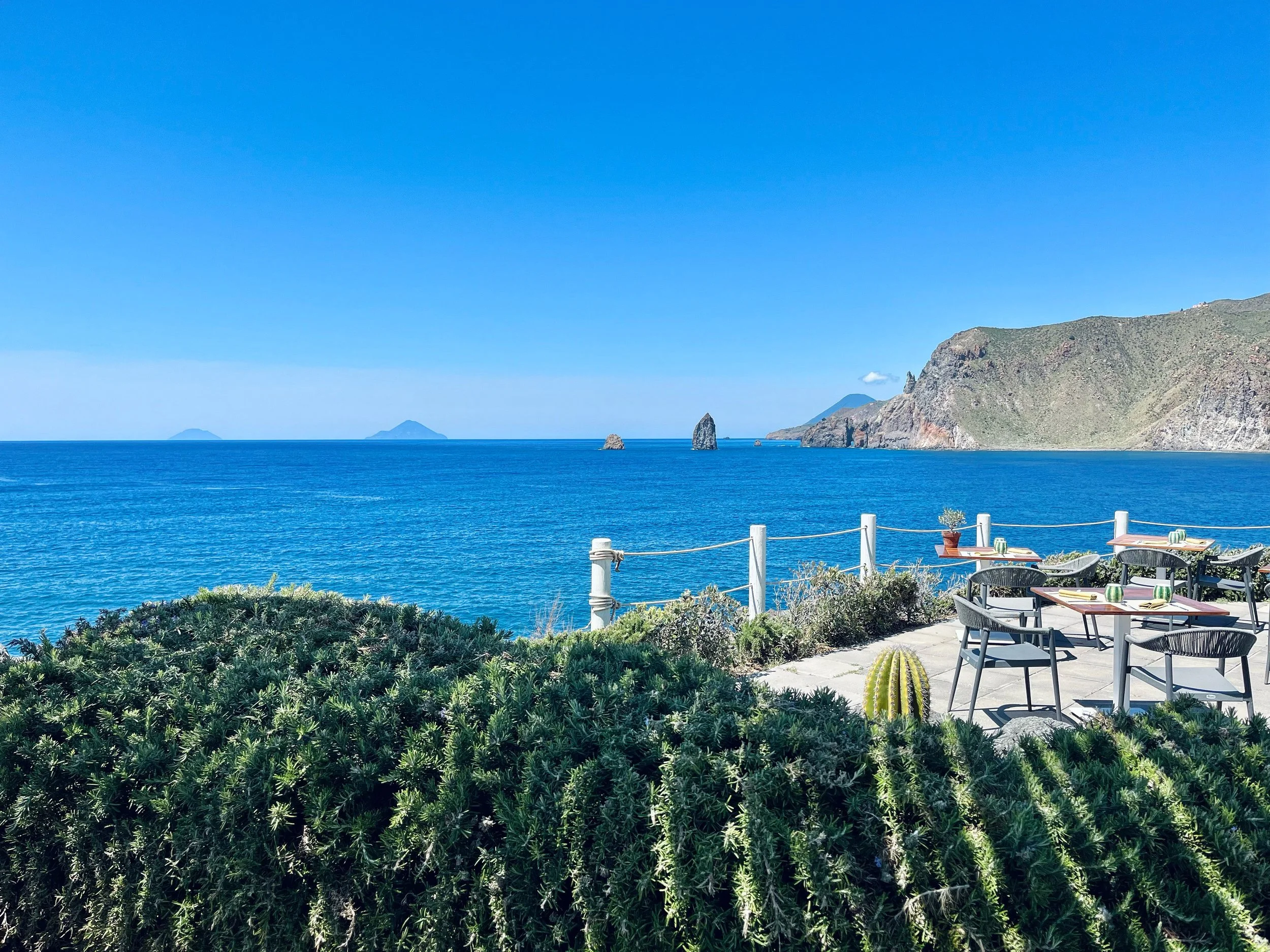 Vista di un'area ristorante all'aperto con tavoli e sedie vicino al mare, con un paesaggio di scogli e isole lontane sotto un cielo azzurro.
