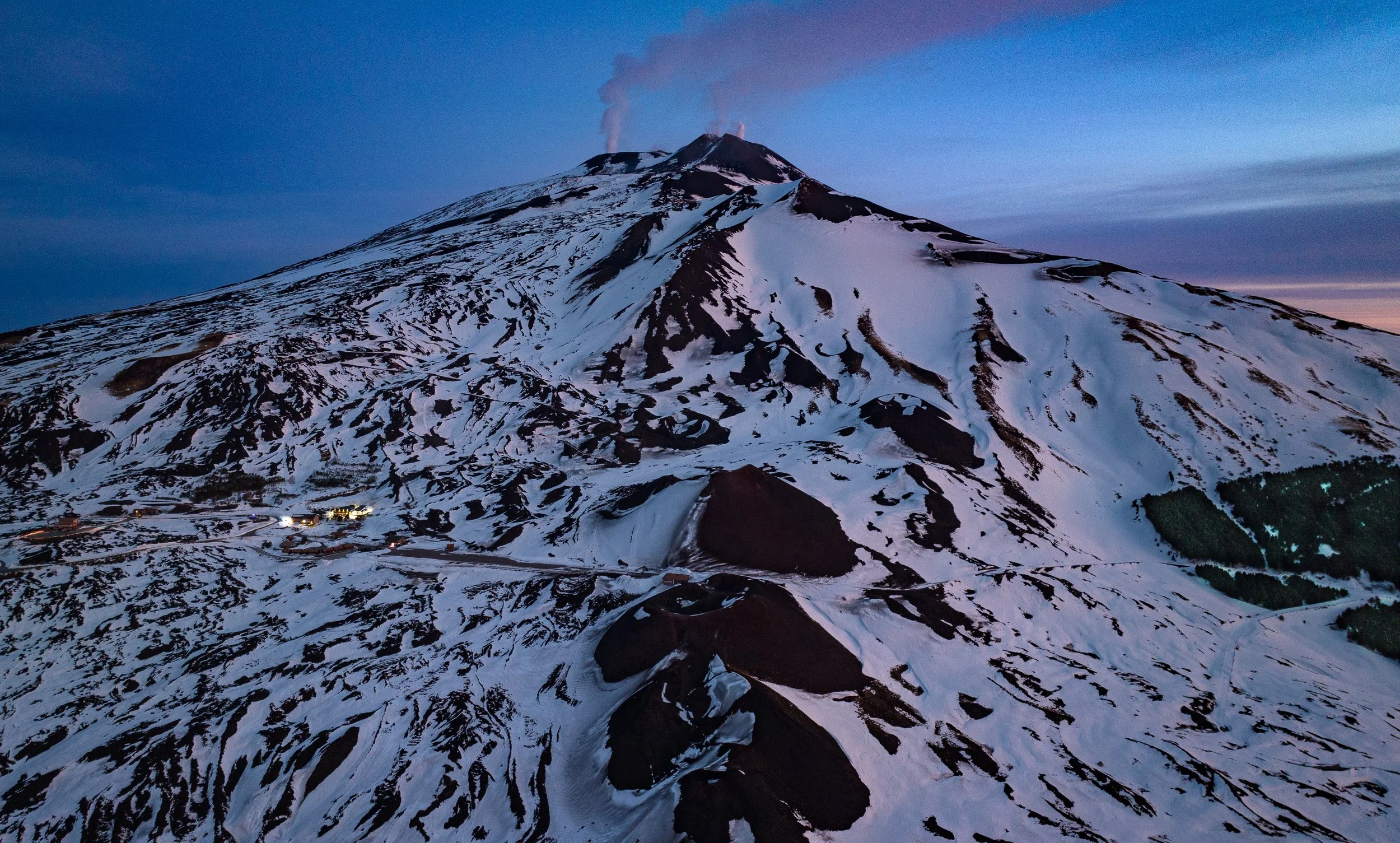 Un vulcano con la cima fumante durante il tramonto, coperto di neve e con un paesaggio montano circostante.