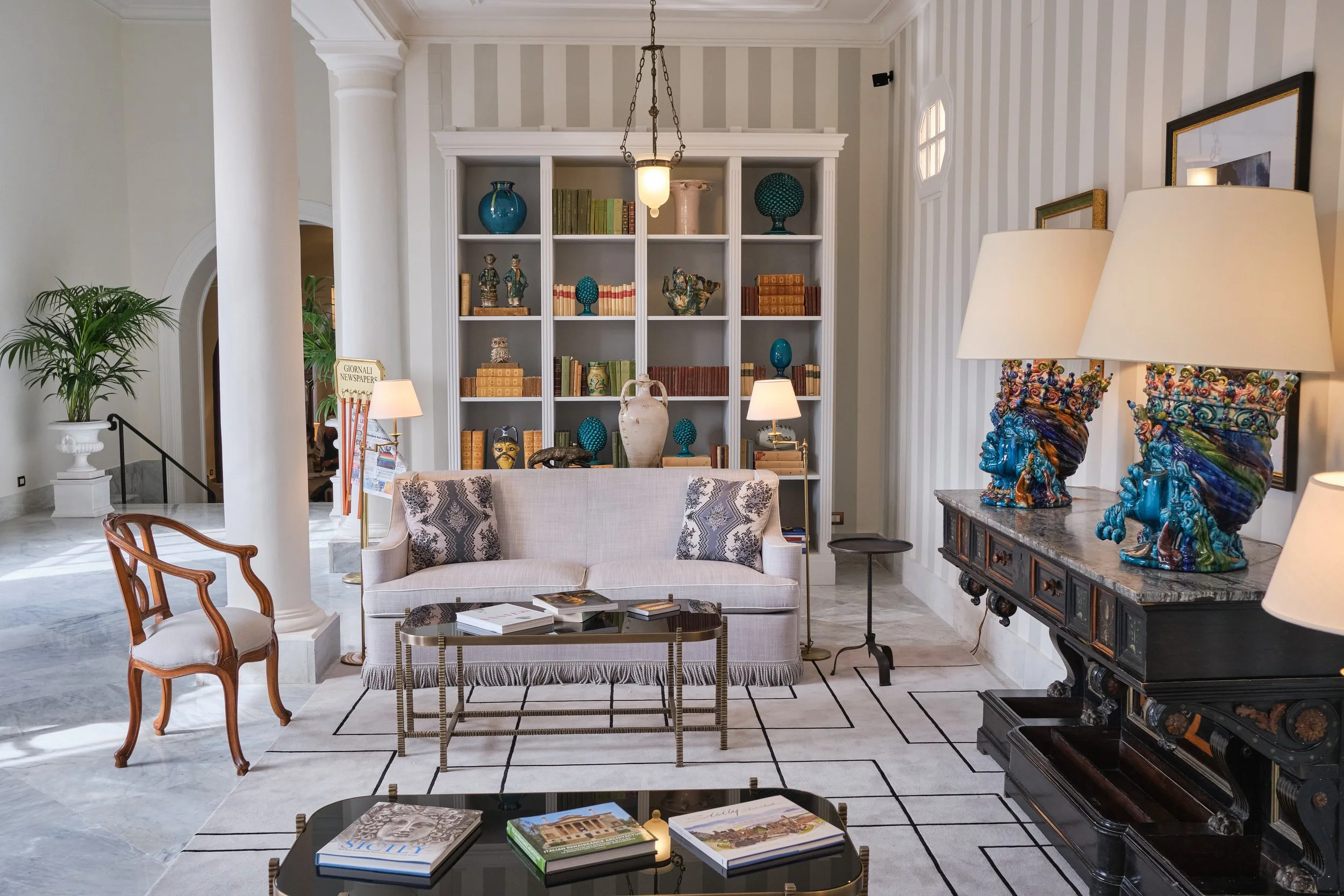 Interno di soggiorno elegante con libreria bianca piena di decorazioni e libri, divano beige con cuscini decorativi, tavolino con riviste, lampade con lampadine, decorazioni di vasi e statuette in vari colori, pareti con strisce di colore neutro, e a