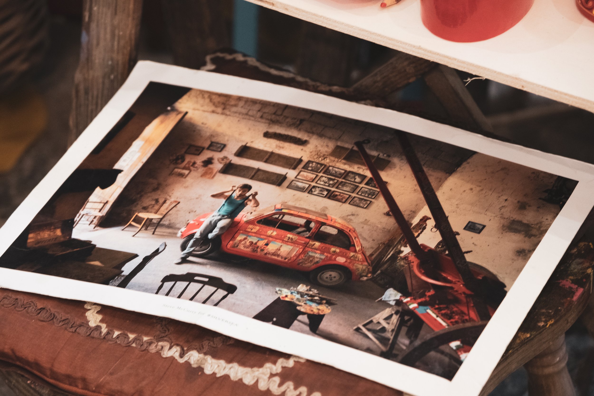 Fotografia di una persona che si riflette in uno specchio, seduta su una sedia in un ambiente decorato con quadri alle pareti e un'auto decorata dipinta con colori vivaci, con scaffali e oggetti artistici sullo sfondo.