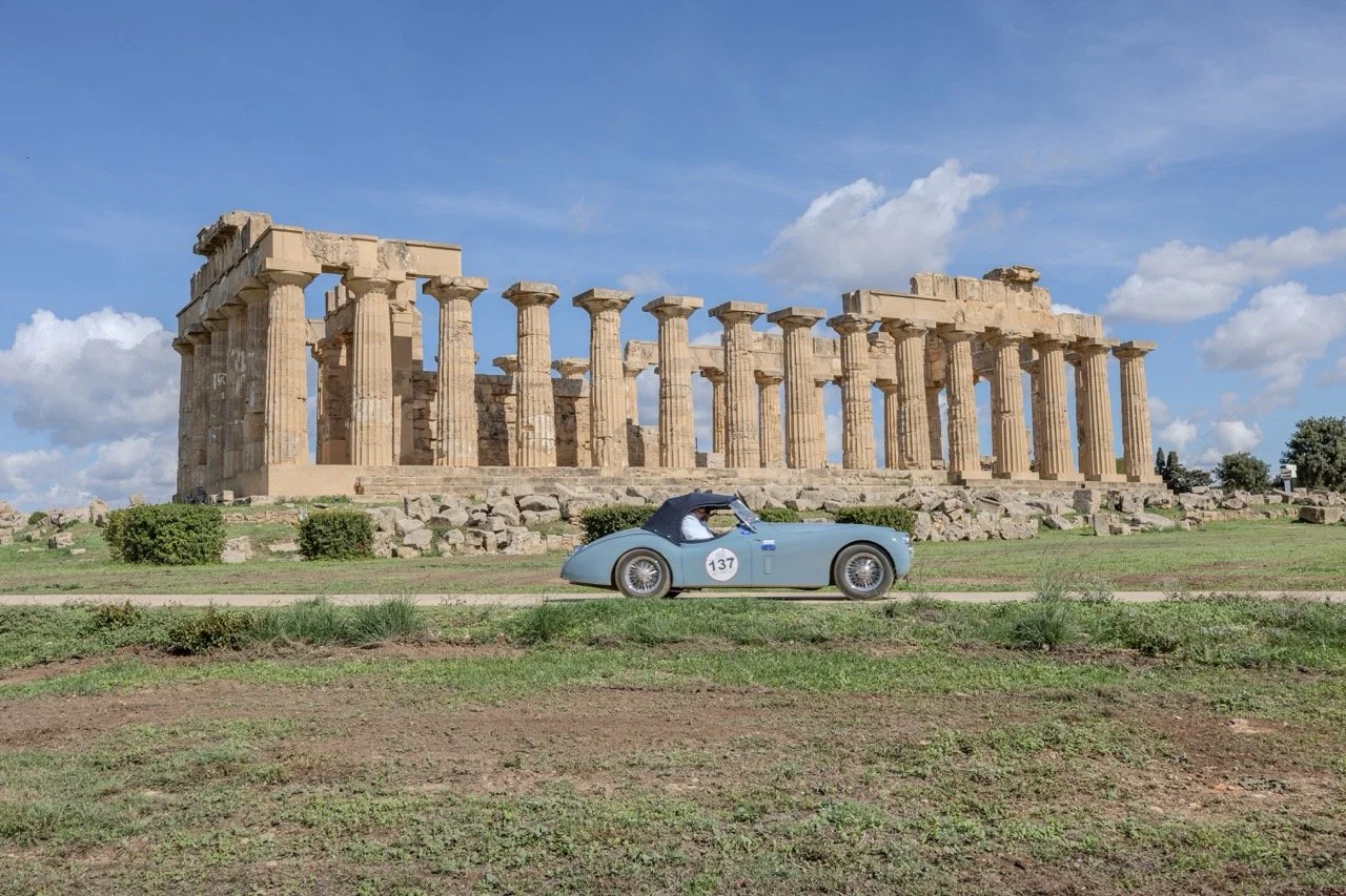 Resti di un tempio antico con colonne doriche, con un'auto da corsa vintage azzurro in primo piano, sotto un cielo con nuvole.