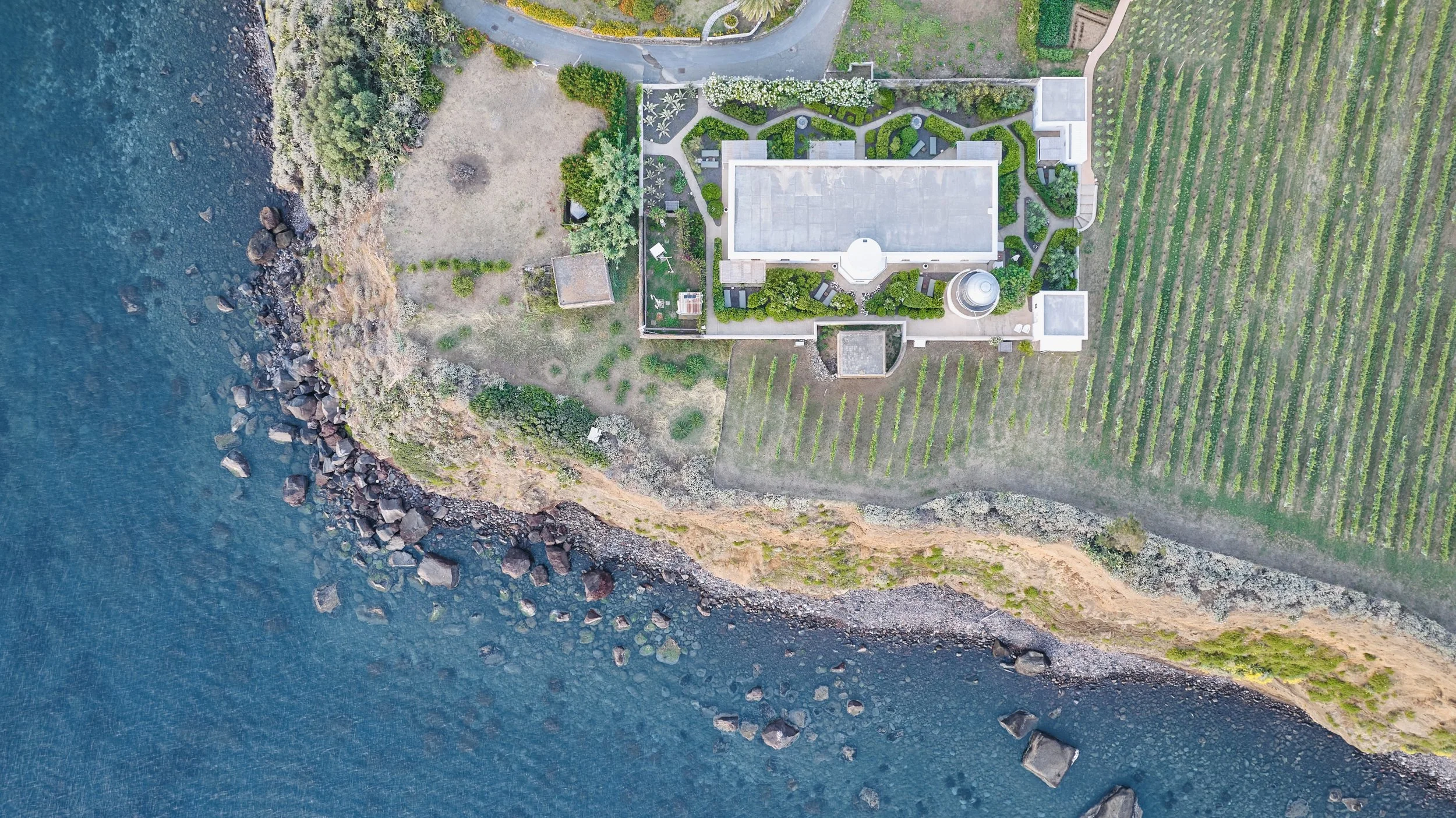 Vista aerea di una casa vicino al mare con giardini ben curati e campi coltivati.