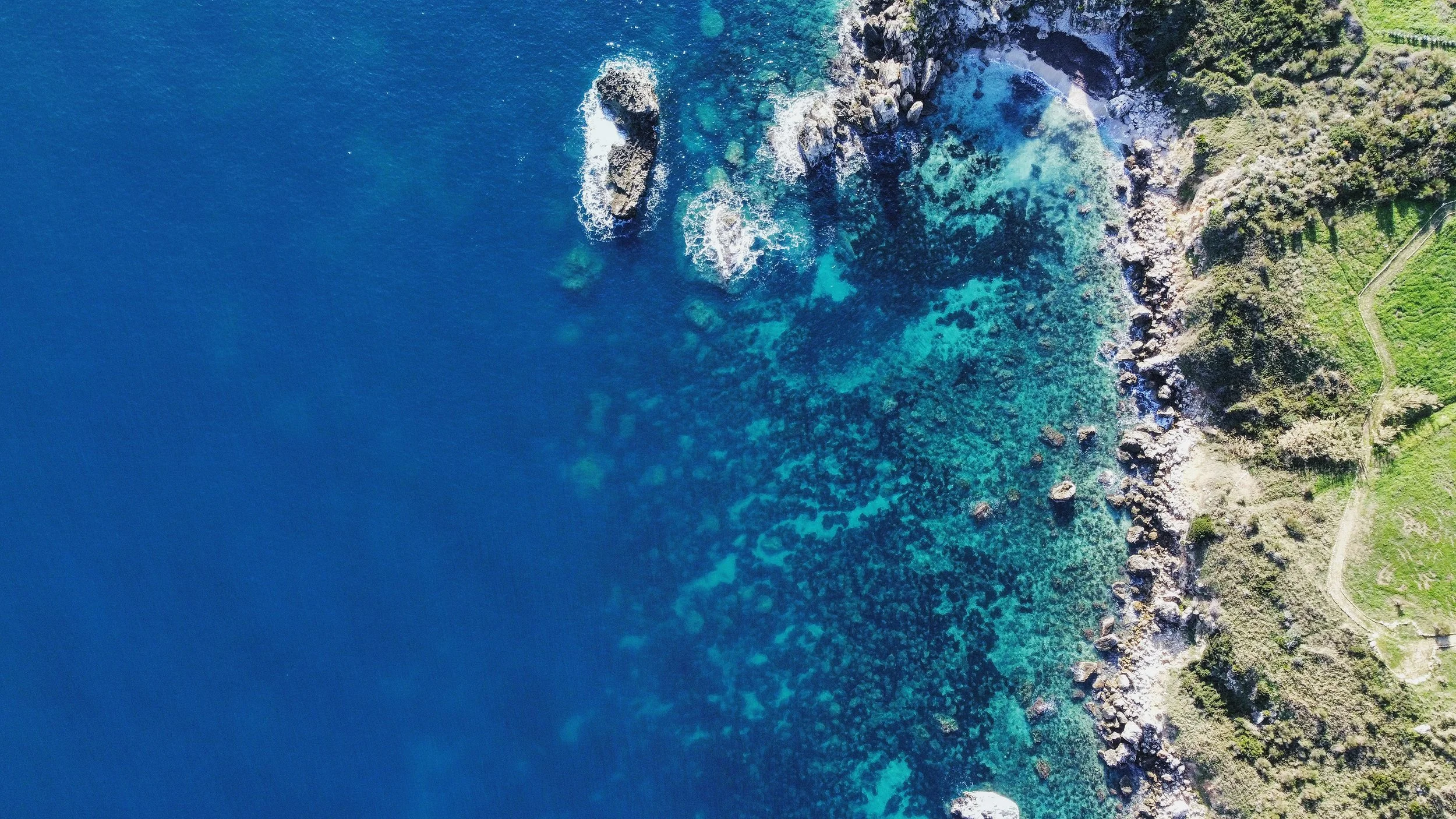 Immagine aerea di una costa con mare blu intenso, acque più chiare vicino alla riva, e una zona verde con vegetazione e sentieri.
