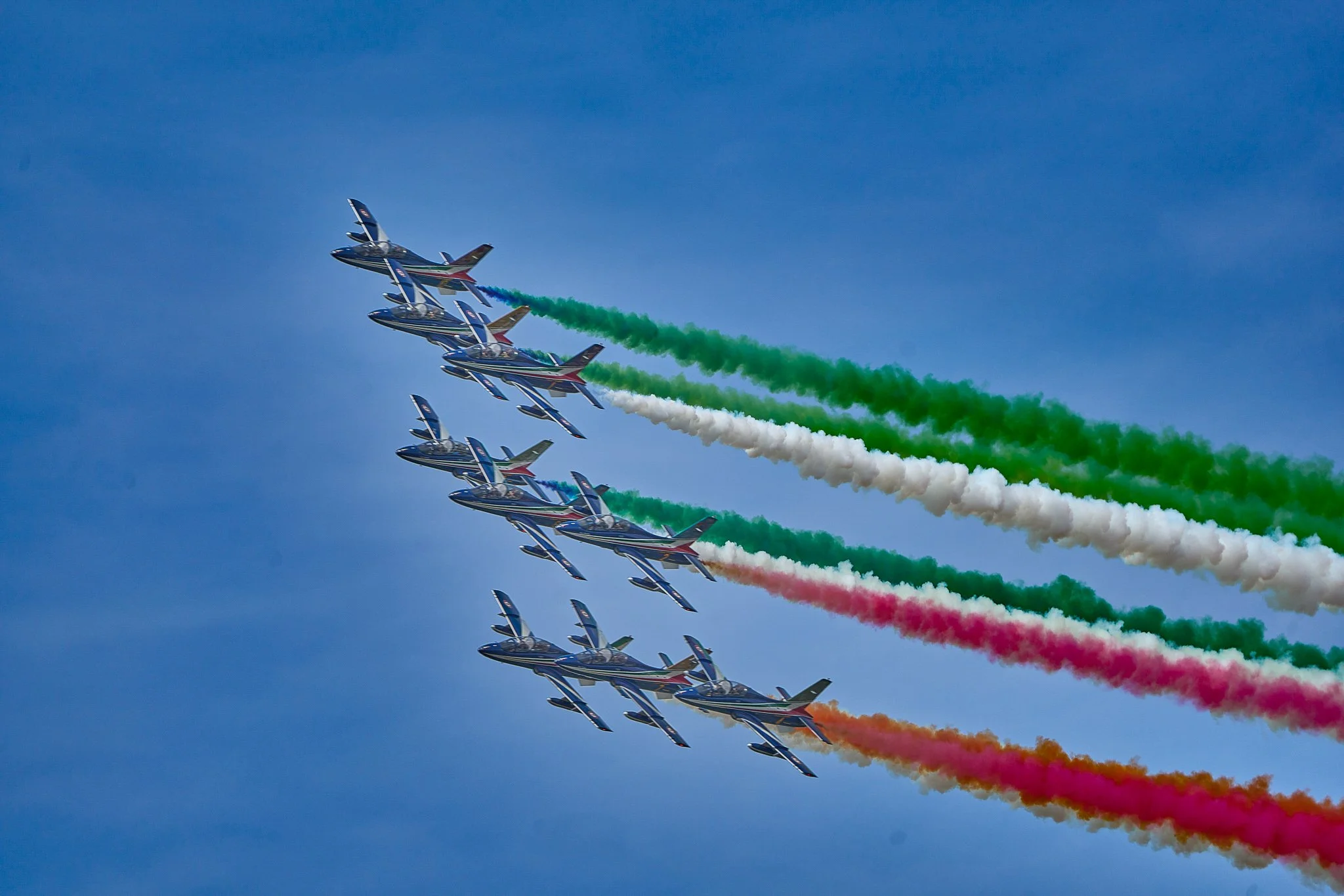 Aerei militari in formazione che rilasciano scie colorate di verde, bianco, rosso e arancione nel cielo blu durante una performance aerea.