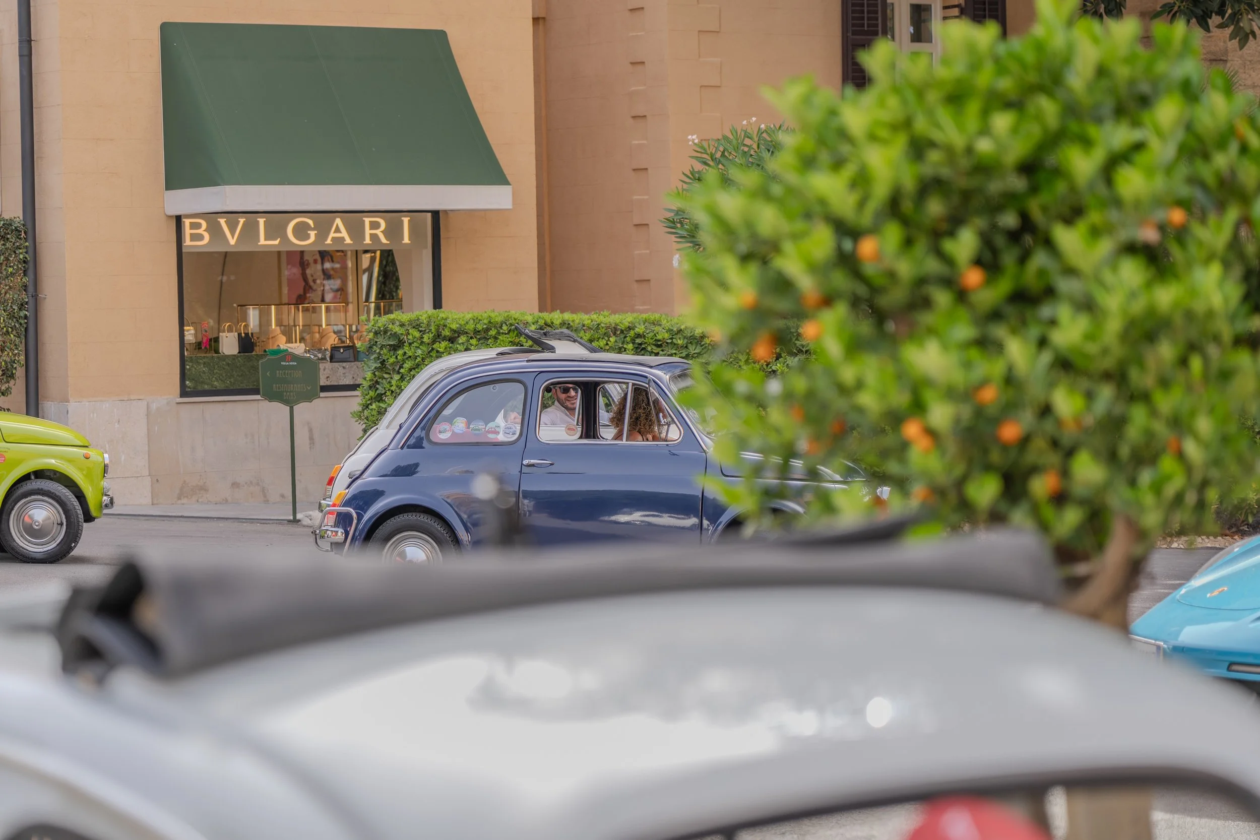 Quattro auto d'epoca parcheggiate davanti a un negozio di lusso con scritta Bvlgari sulla facciata, con alcune persone sedute all'interno di una delle auto, vista parziale di un ambiente urbano con edifici in pietra e vegetazione.