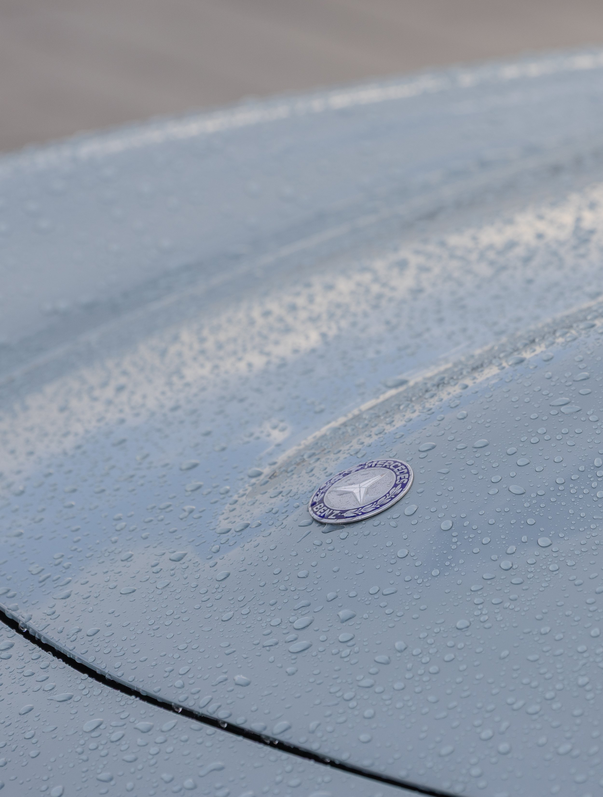 Dettaglio di una superficie di un'auto Mercedes-Benz metallizzata con goccioline di pioggia e un nostro logo sul cofano.