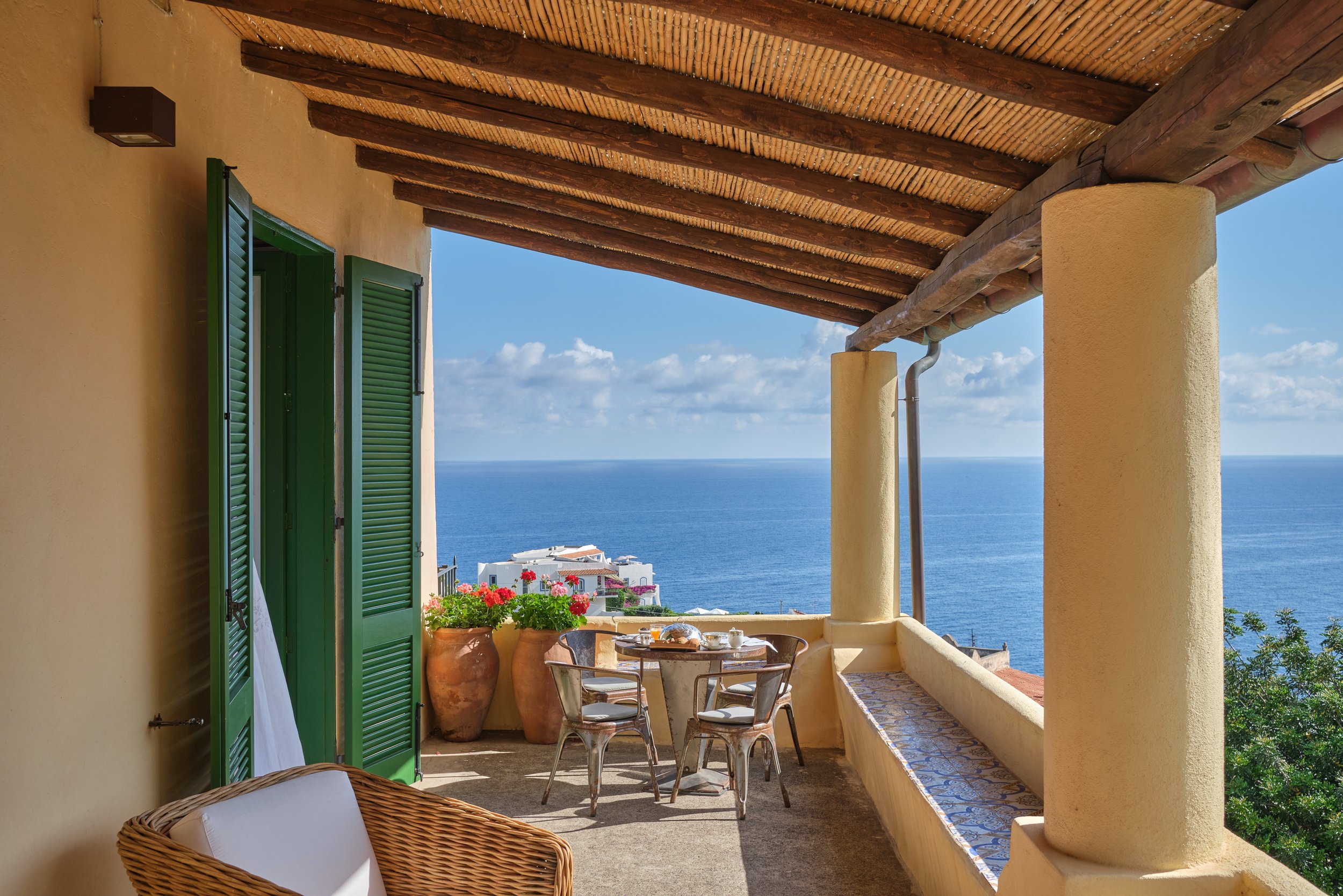 Balcone di una casa con vista sul mare, decorato con fiori in vasi di terracotta, tavolo e sedie di metallo, colonne e tetto in legno