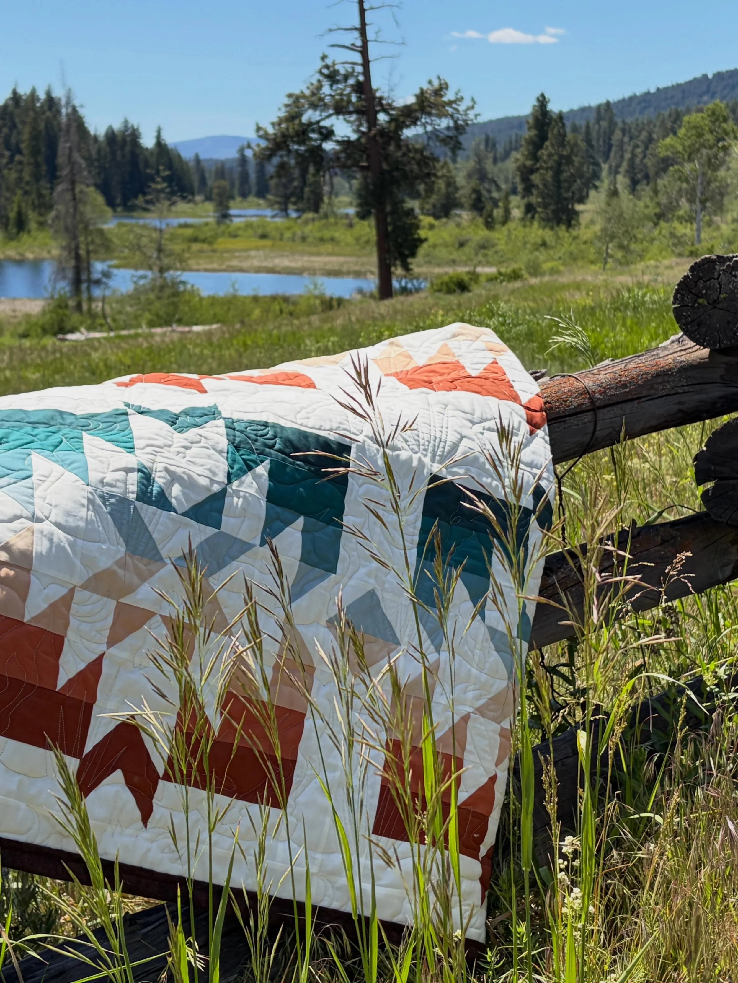 A quilted blanket with geometric patterns in teal, white, orange, and brown is draped over a fallen tree log, surrounded by tall grass. In the background, there is a landscape with a river, green trees, hills, and mountains under a blue sky with some clouds.