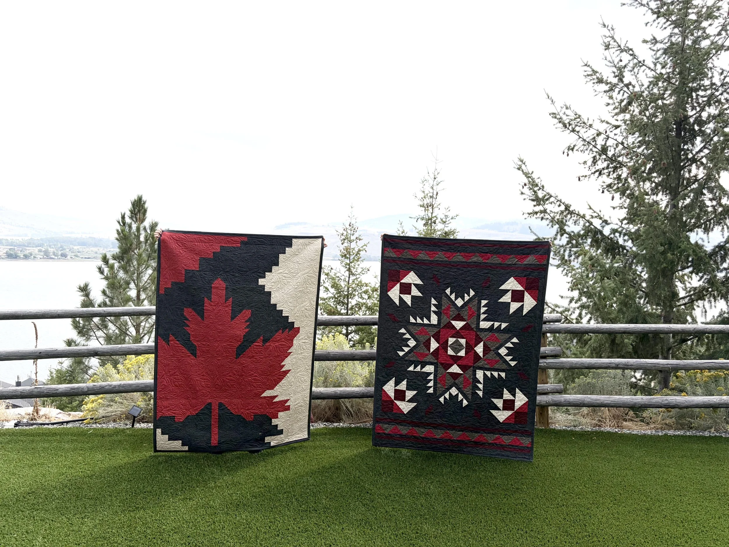 Two quilts with Native American-inspired geometric patterns hanging on a wooden railing outdoors, with trees, a lake, and mountains in the background.