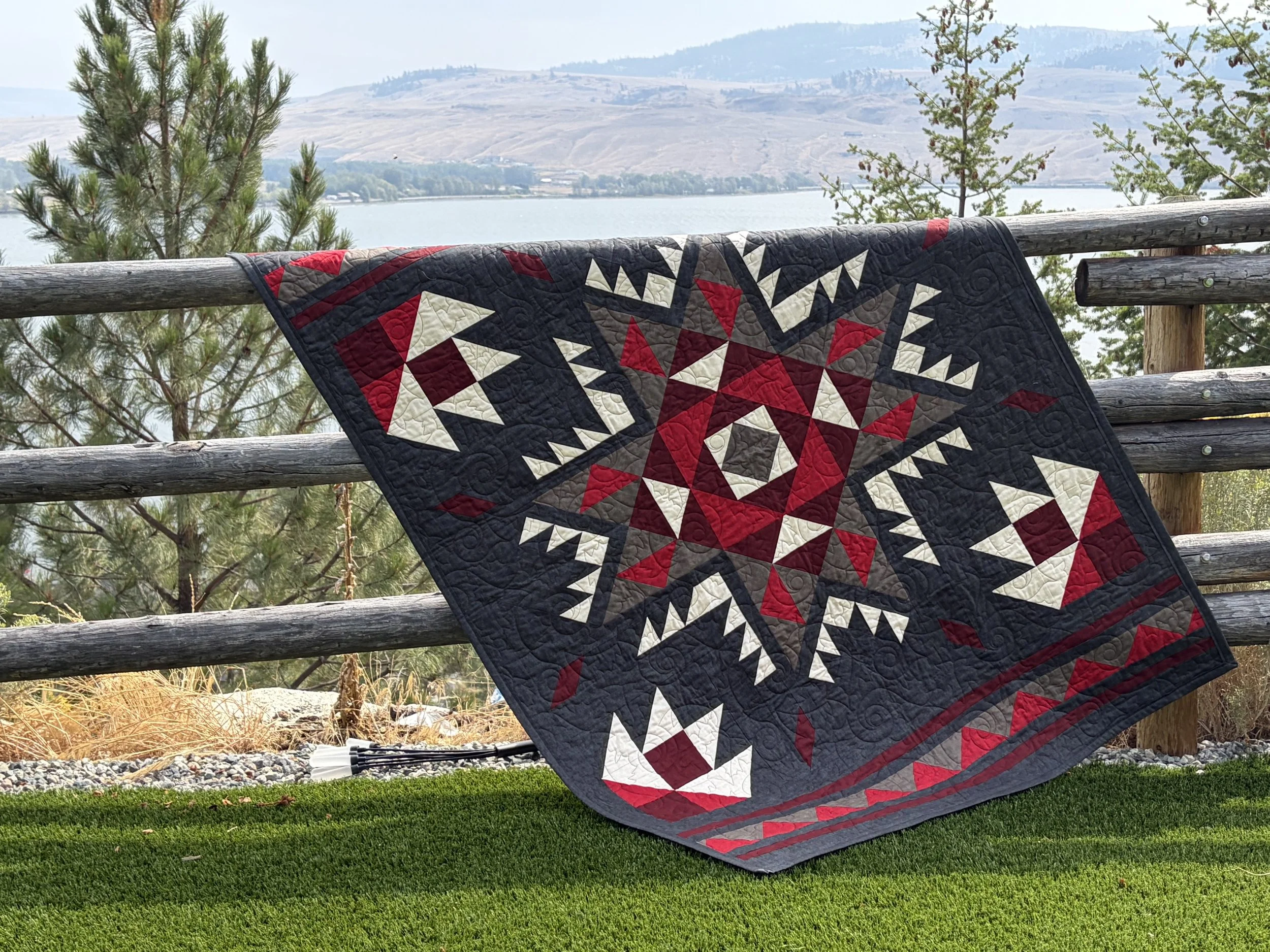 A quilt with a red, black, and white geometric pattern is draped over a wooden railing on an outdoor deck, overlooking a lake with trees and rolling hills in the background.