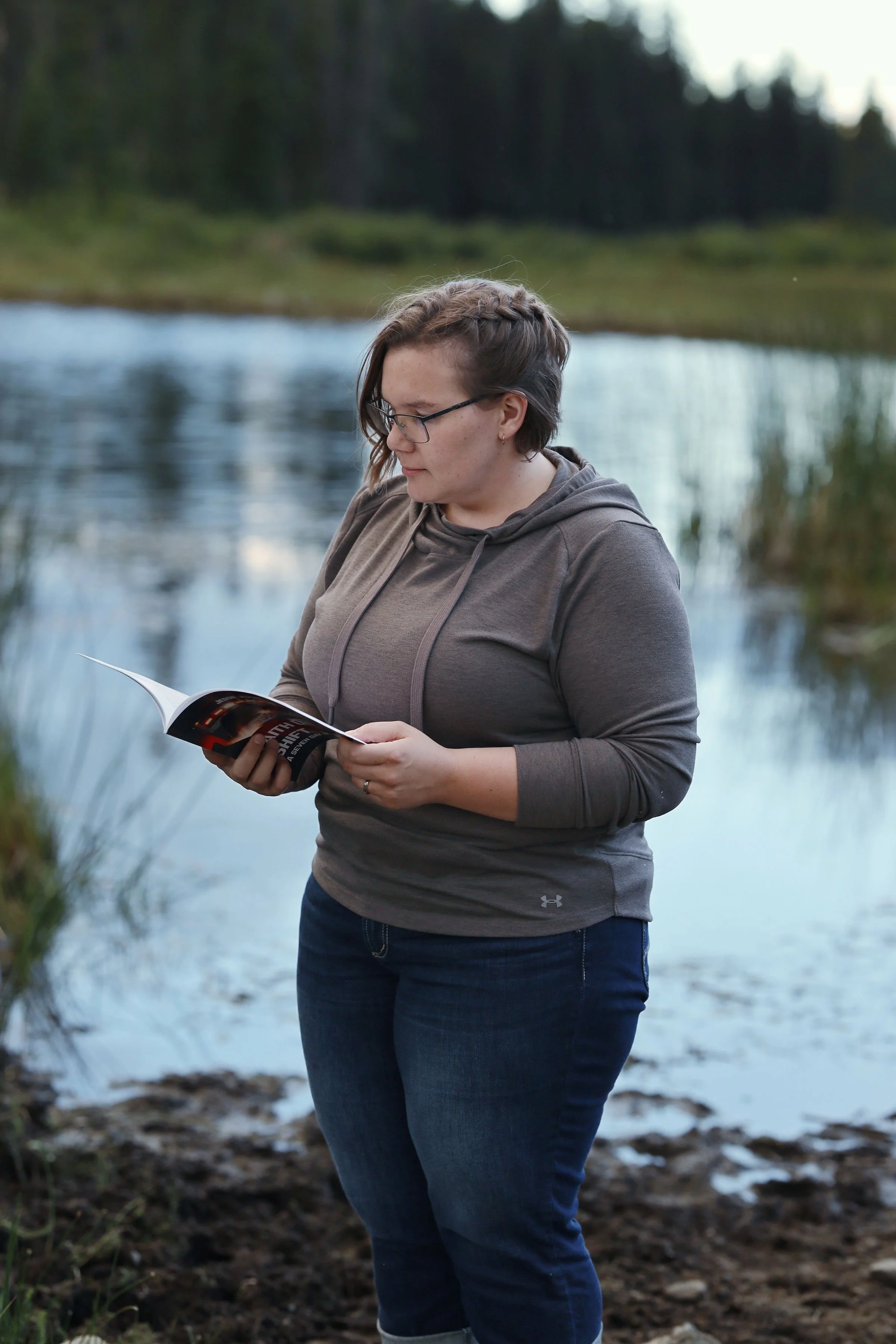 A woman with short hair, glasses, and a gray hoodie stands near a body of water, reading a magazine outdoors during daytime.