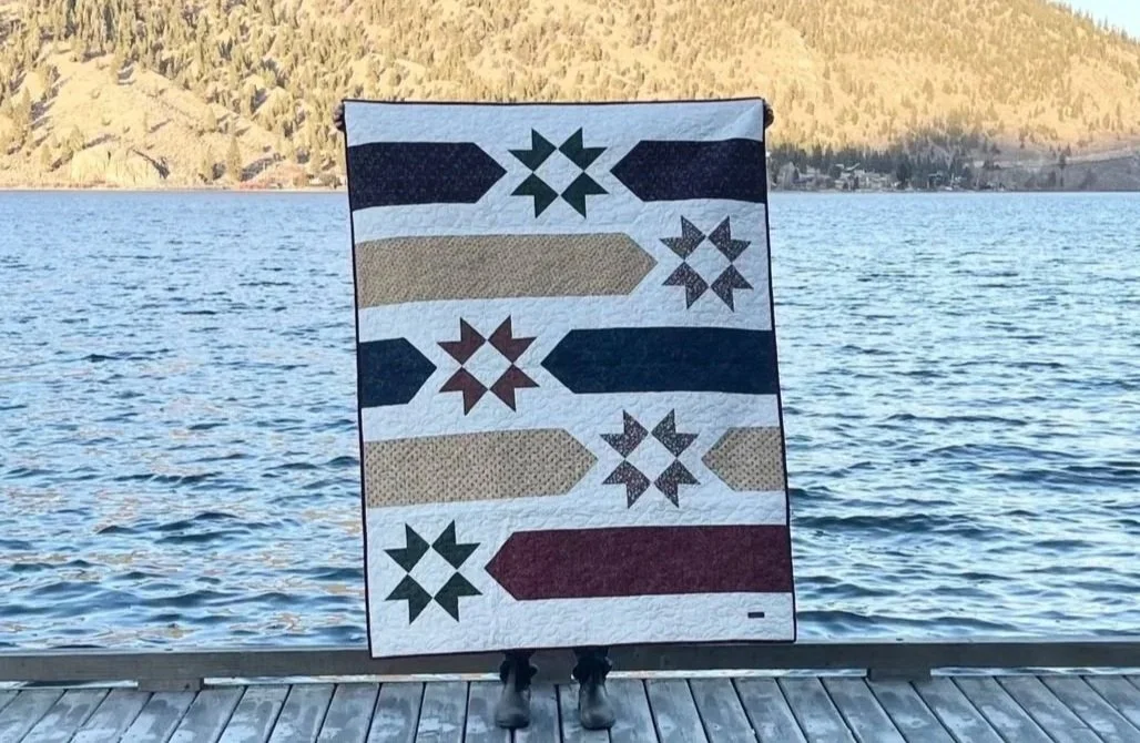 Person holding a quilt with arrow patterns in front of a lake and mountainous landscape.