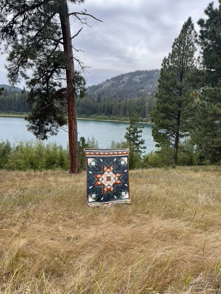 A quilt with a star pattern hanging in a grassy field near a lake with pine trees and mountains in the background.