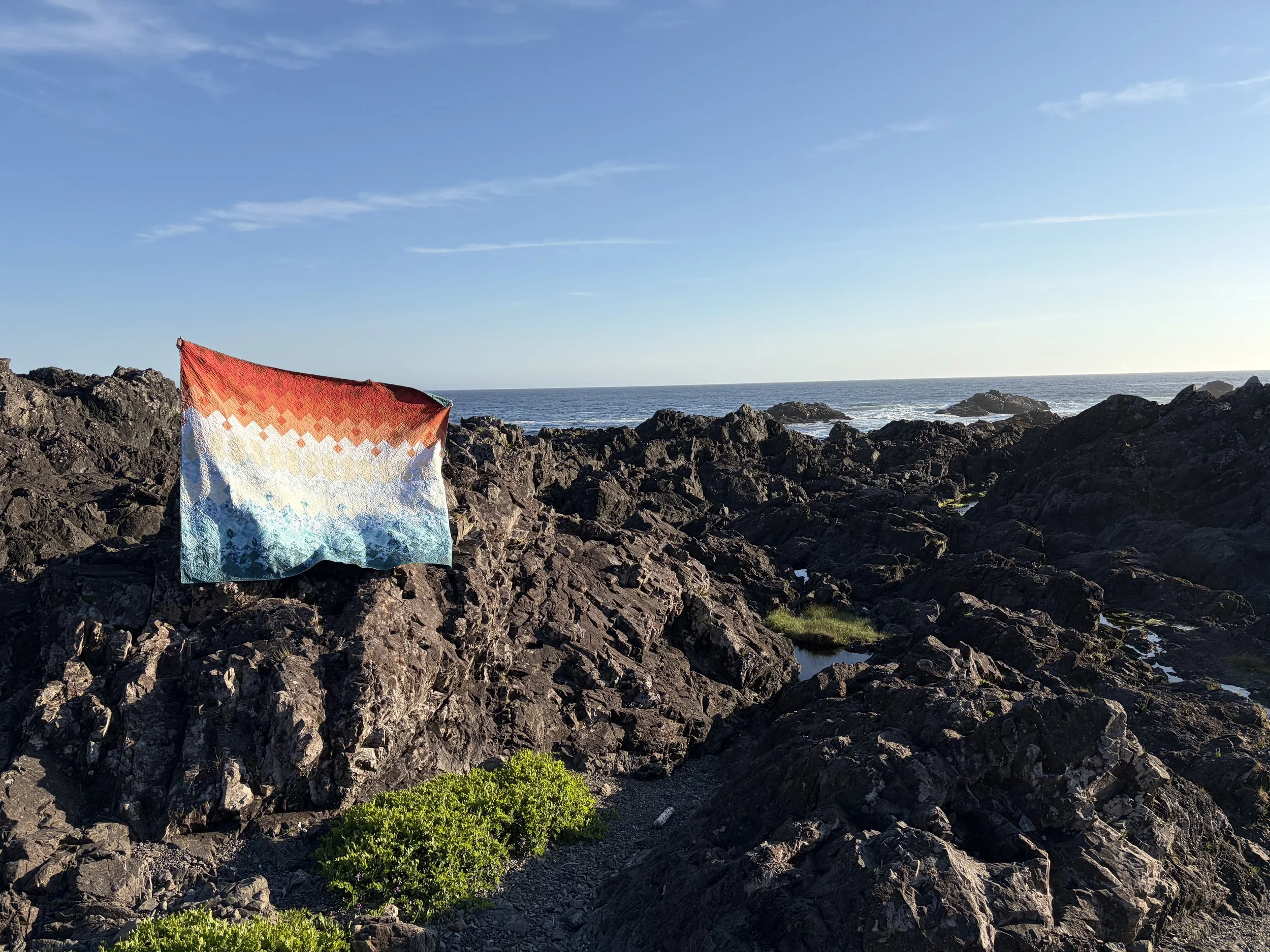 A rocky coastline with sparse green vegetation, a colorful patterned blanket draped over rocks, and the ocean with a clear blue sky in the background.