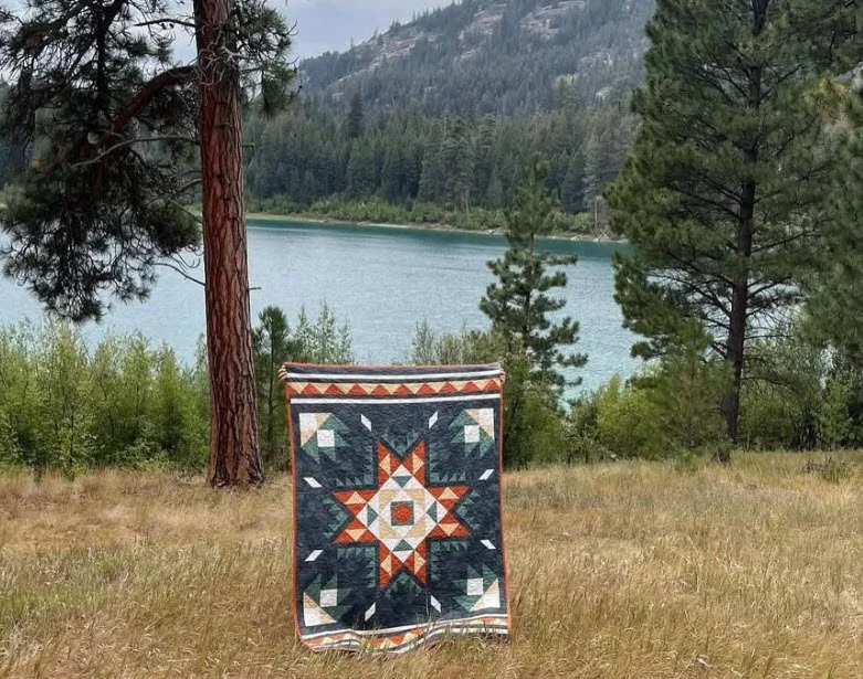 A quilt with a geometric star pattern is draped over a foreground in front of a lake, with pine trees and mountains in the background.