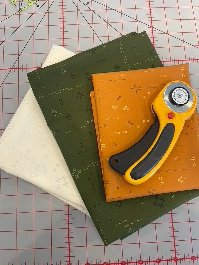 Four folded pieces of fabric in white, dark green, and orange colors, stacked on a cutting mat. A rotary cutter with a yellow handle is resting on top of the orange fabric.