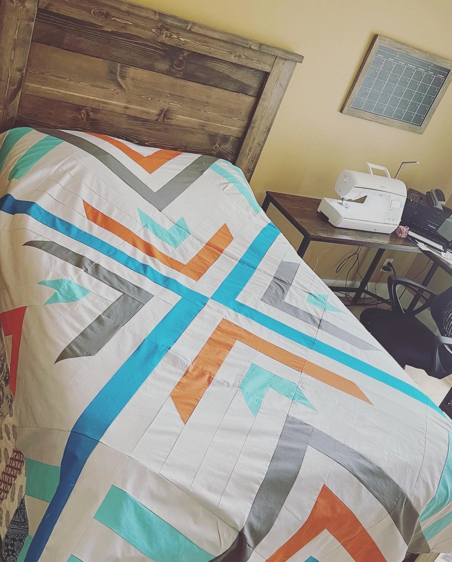 A colorful quilt with geometric patterns on a bed with a wooden headboard in a bedroom, next to a desk with a sewing machine, a printer, and a black chair.